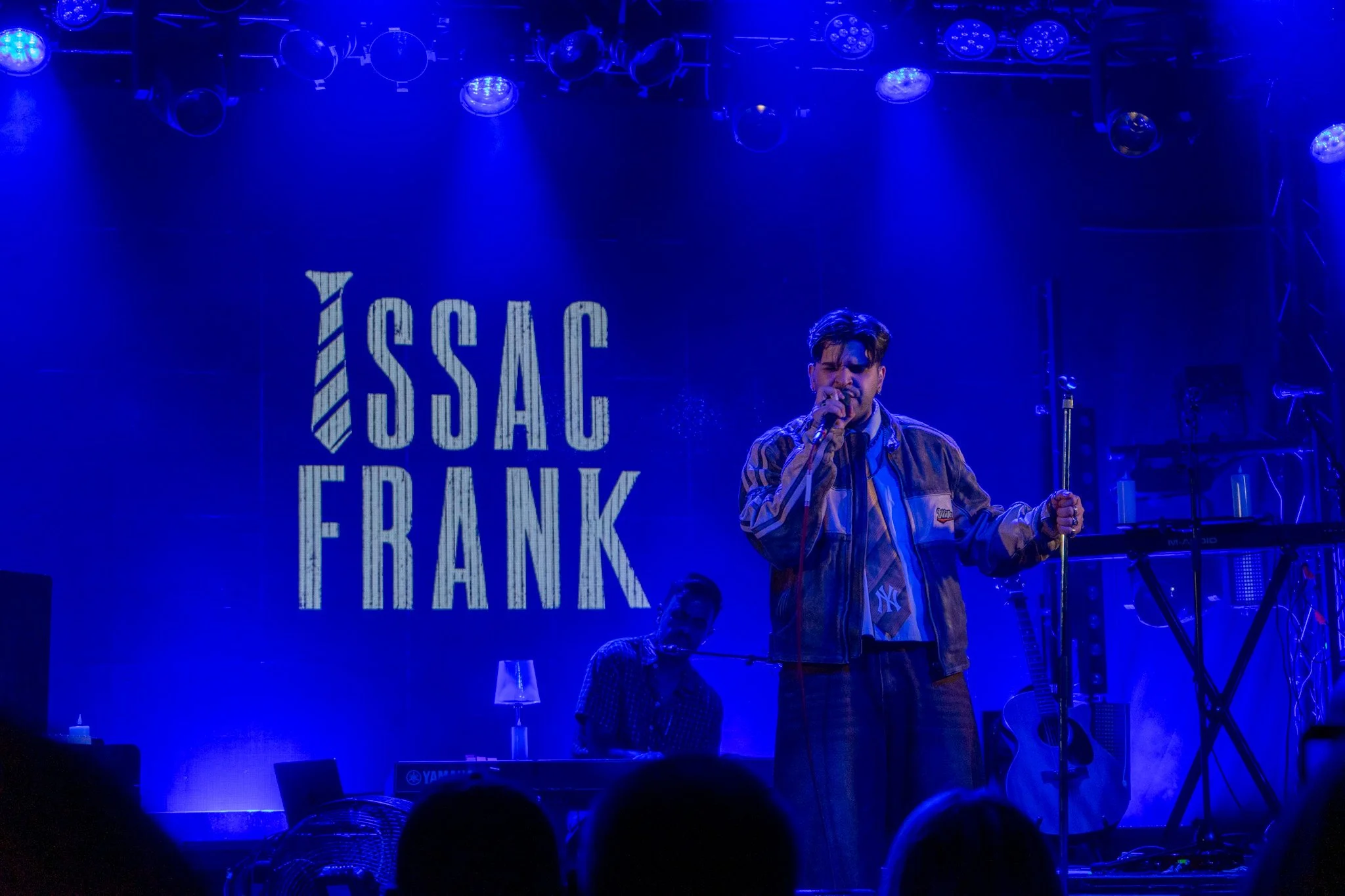 A musical performance on a stage with blue lighting, featuring a singer holding a microphone. Behind him, a large screen displays the text 'YASSO' and 'REAK'. There are other musicians playing instruments on stage.