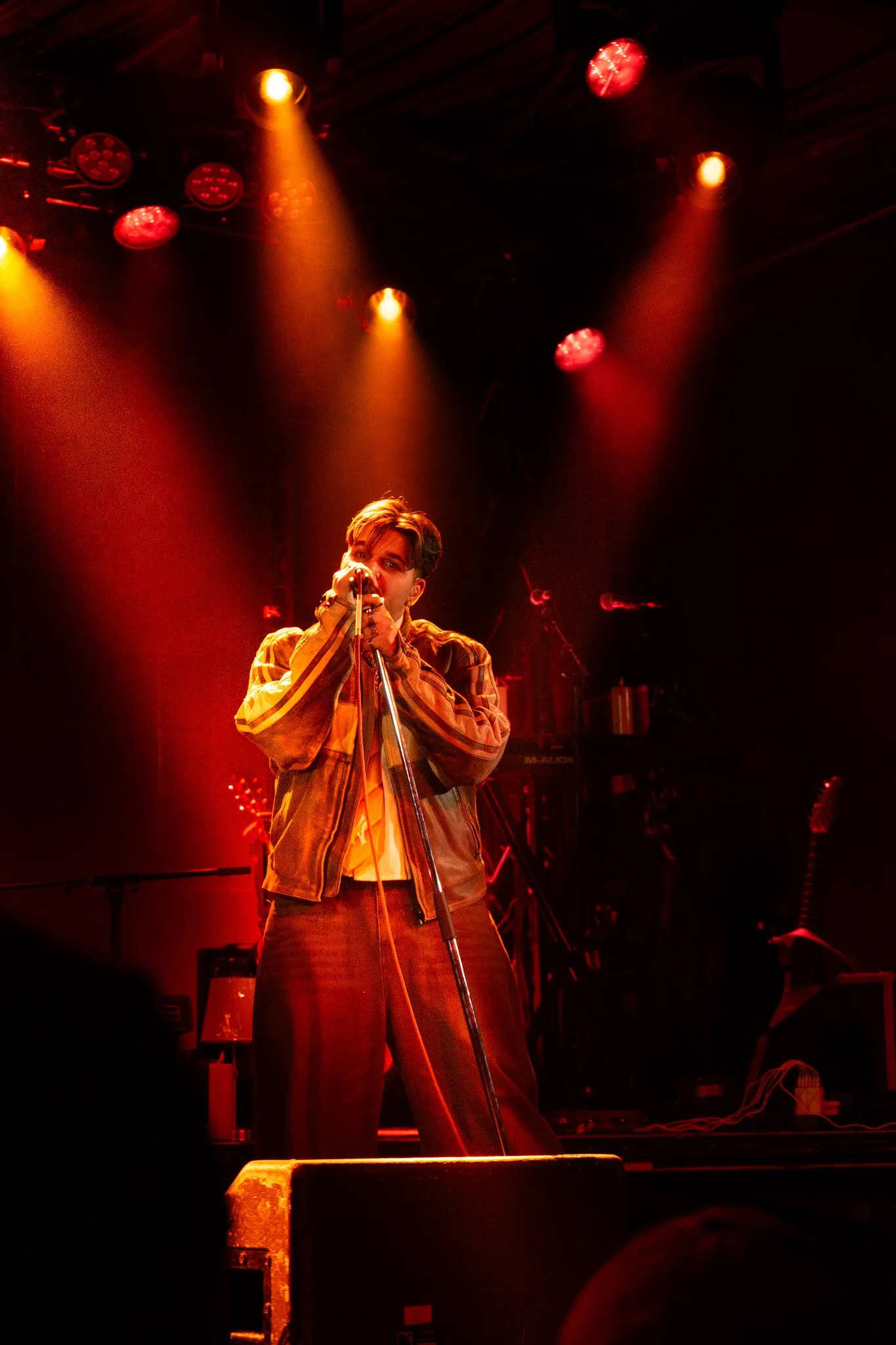 A young male singer wearing an orange and yellow striped jacket performing on stage with red spotlighting and musical instruments in the background.