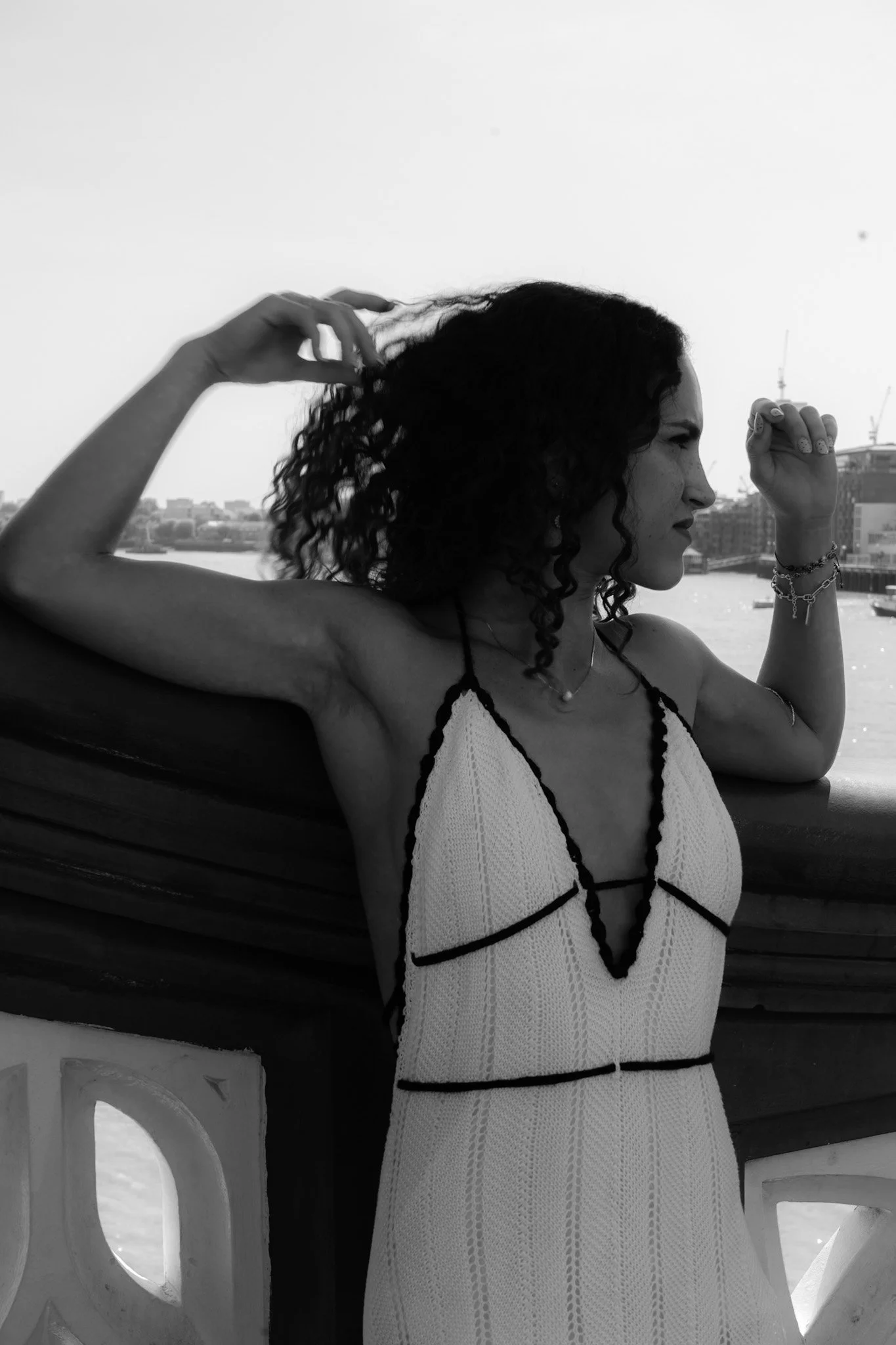A woman with curly hair is standing outdoors near a body of water, adjusting her hair with her right hand, wearing a white dress with black trim, jewelry, and bracelets.