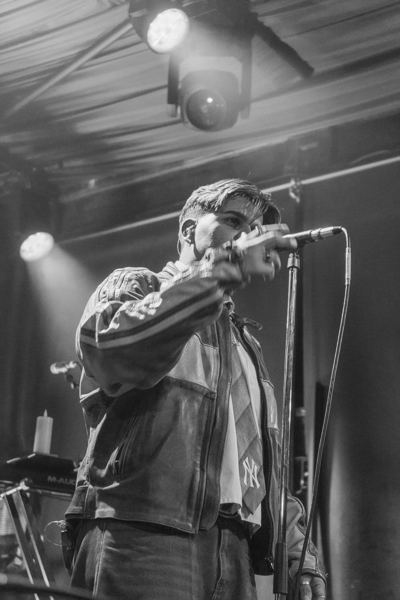 Black and white photo of a young man wearing a jacket, standing behind a microphone, in what appears to be a music or performance studio with stage lights overhead.