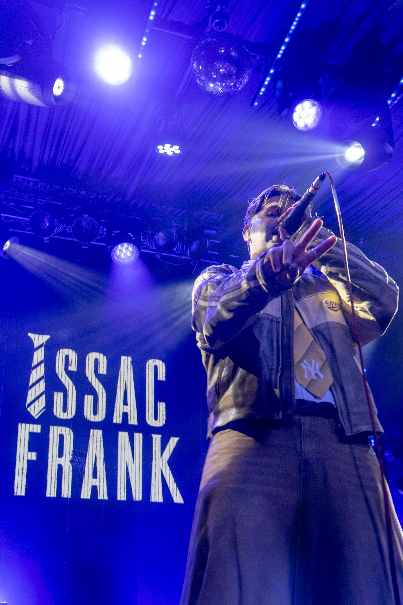 A person singing into a microphone on stage with blue lighting, wearing a plaid jacket and brown pants, with a sign in the background reading 'MISSISSIPPI FRANK'.