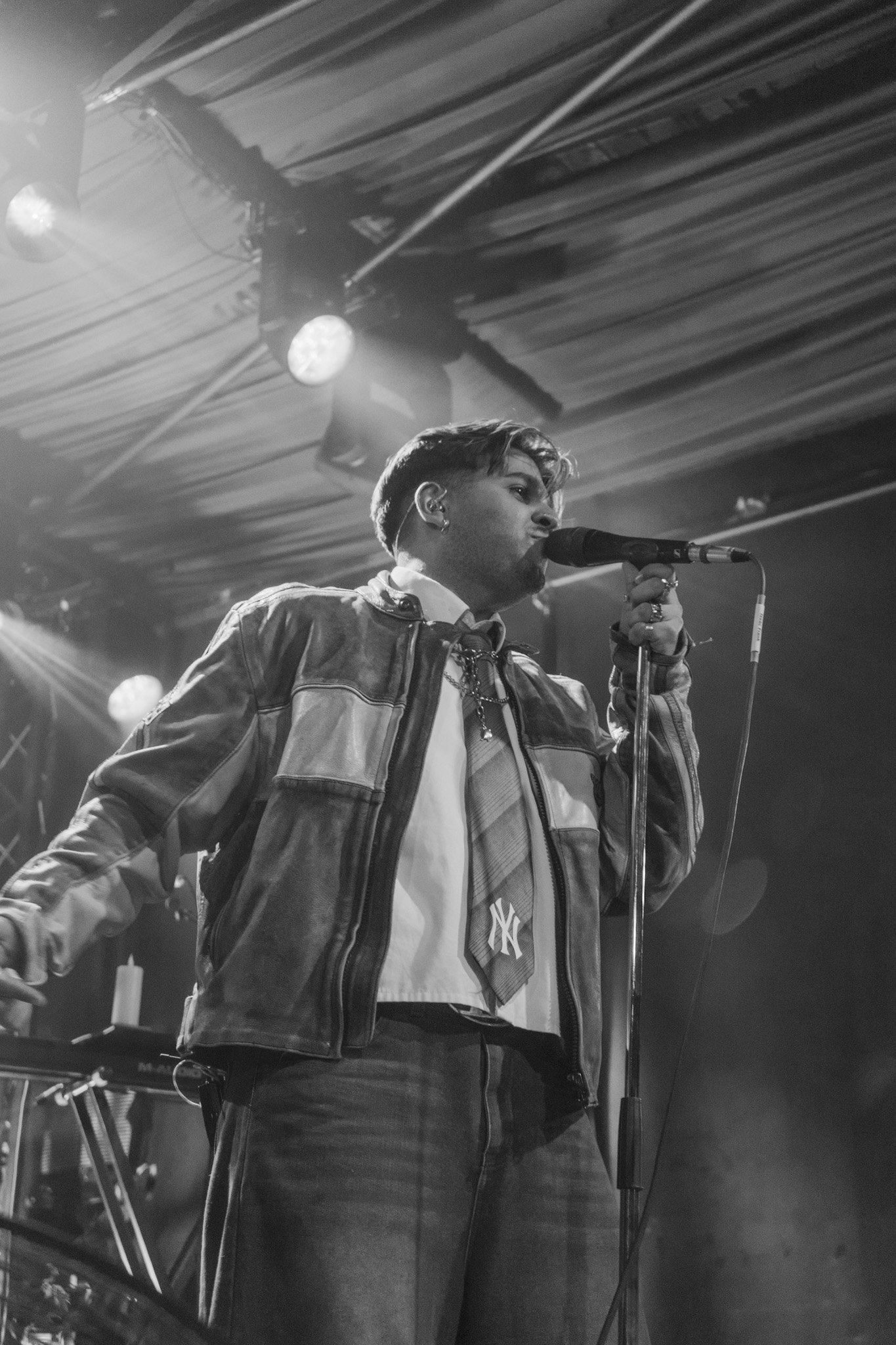 Black and white photo of a young person with short hair and earrings singing into a microphone on stage, wearing a jacket and a shirt with a New York Yankees logo, with stage lights and musical equipment in the background.
