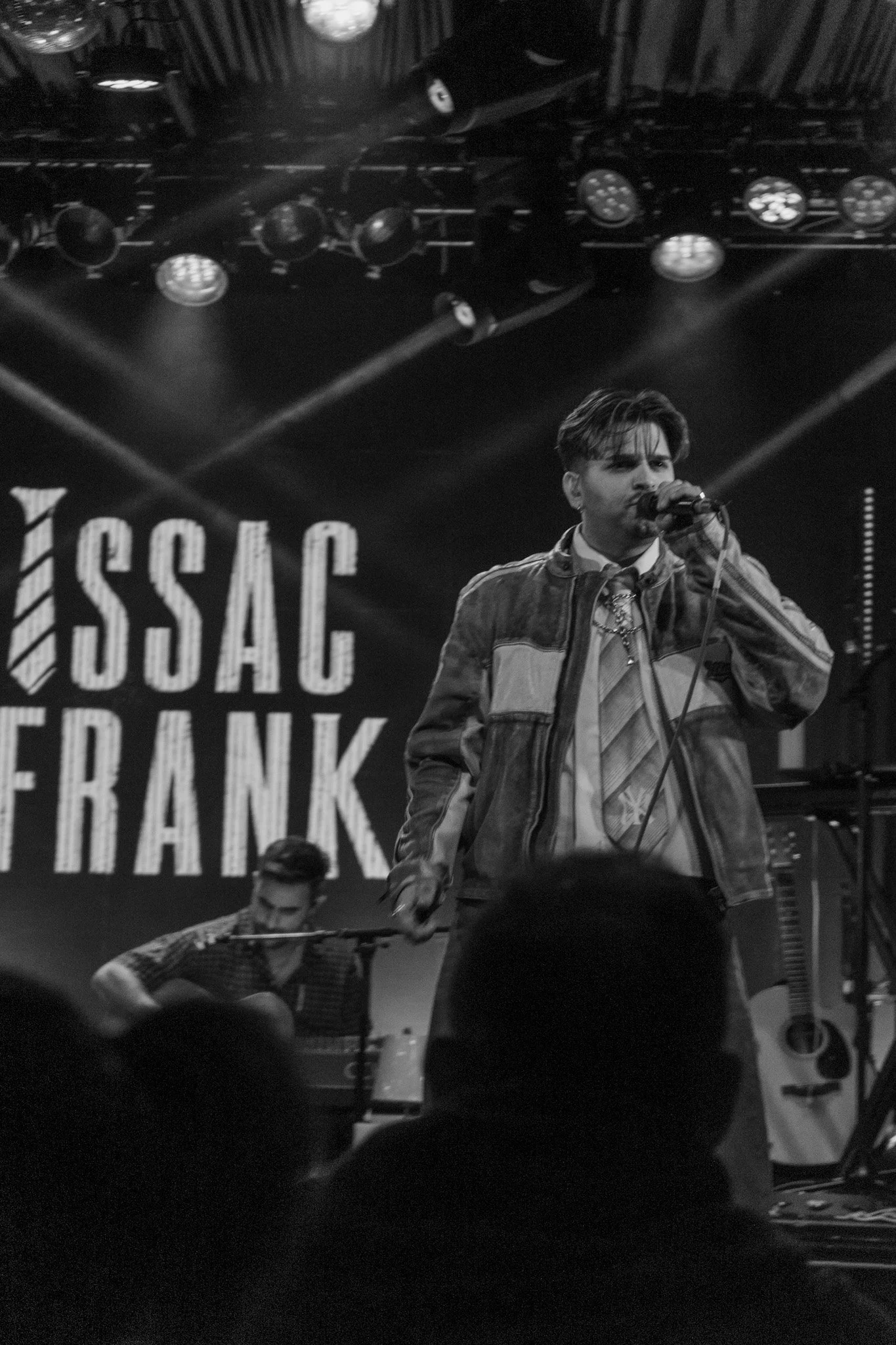 Black and white photo of a man singing into a microphone on stage with a guitarist playing in the background. There is a large sign that reads "ESC" and "BANK" in the background. Stage lighting is visible above.