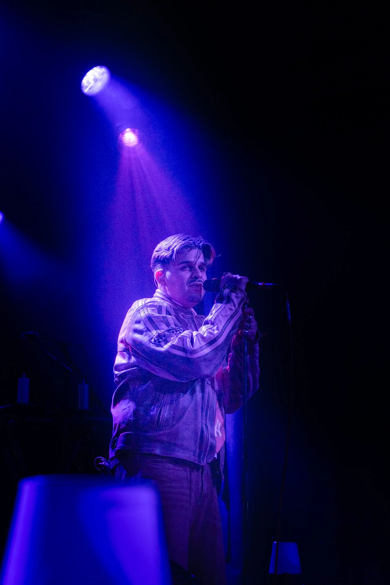 A young man with styled hair and a plaid jacket singing into a microphone on a dark stage illuminated by blue and purple stage lights.