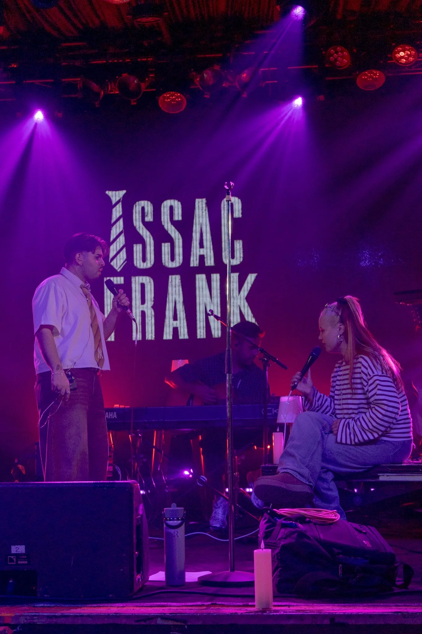 Stage with purple lighting, a man holding a microphone, a woman sitting and speaking into a microphone, and a large screen in the background displaying the words 'ISSAC FRANK'.