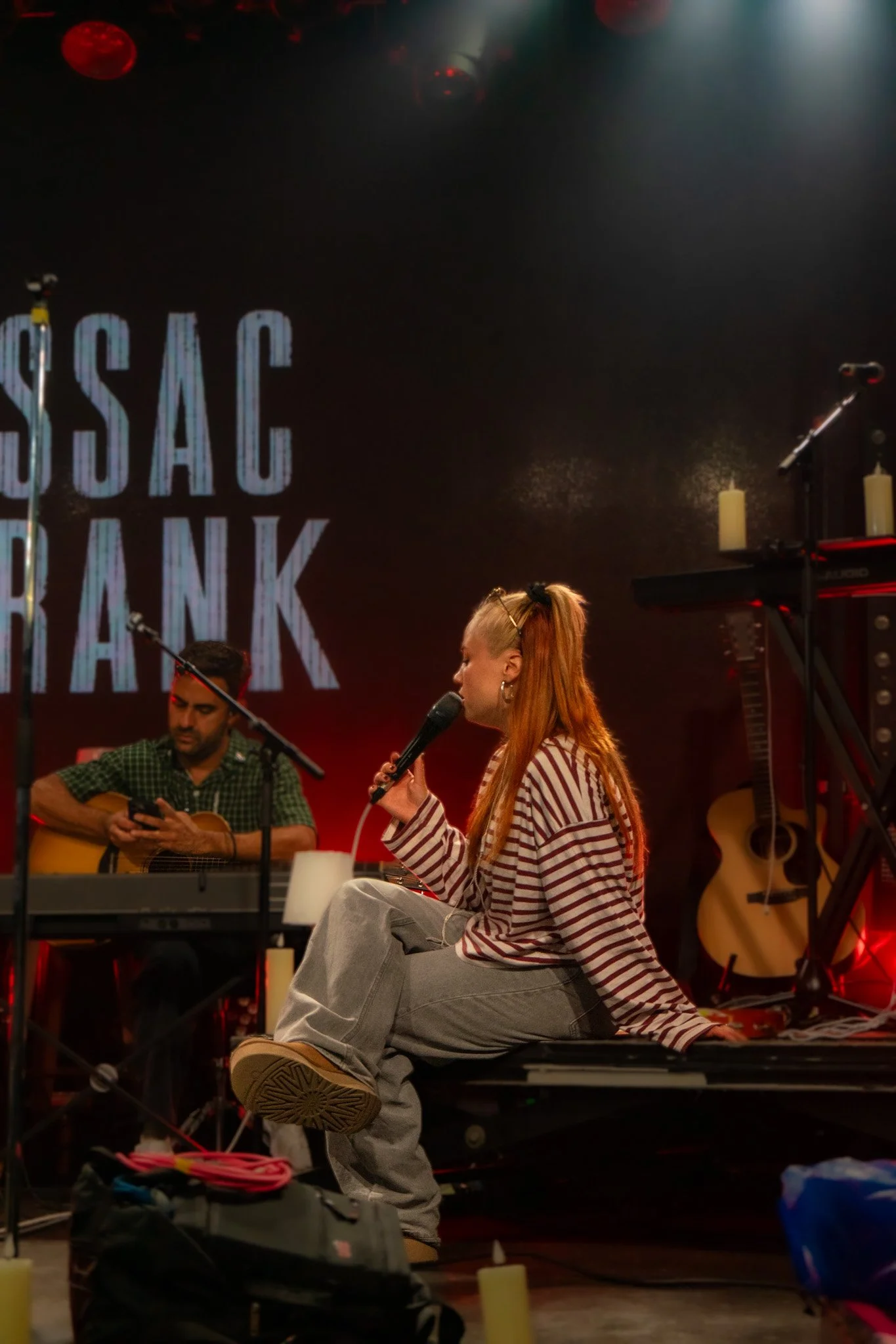 A woman with red hair, wearing a striped red and white shirt and gray sweatpants, sits cross-legged on a stage, holding a microphone. A man with short dark hair and a green checkered shirt plays an acoustic guitar beside her. The background features 