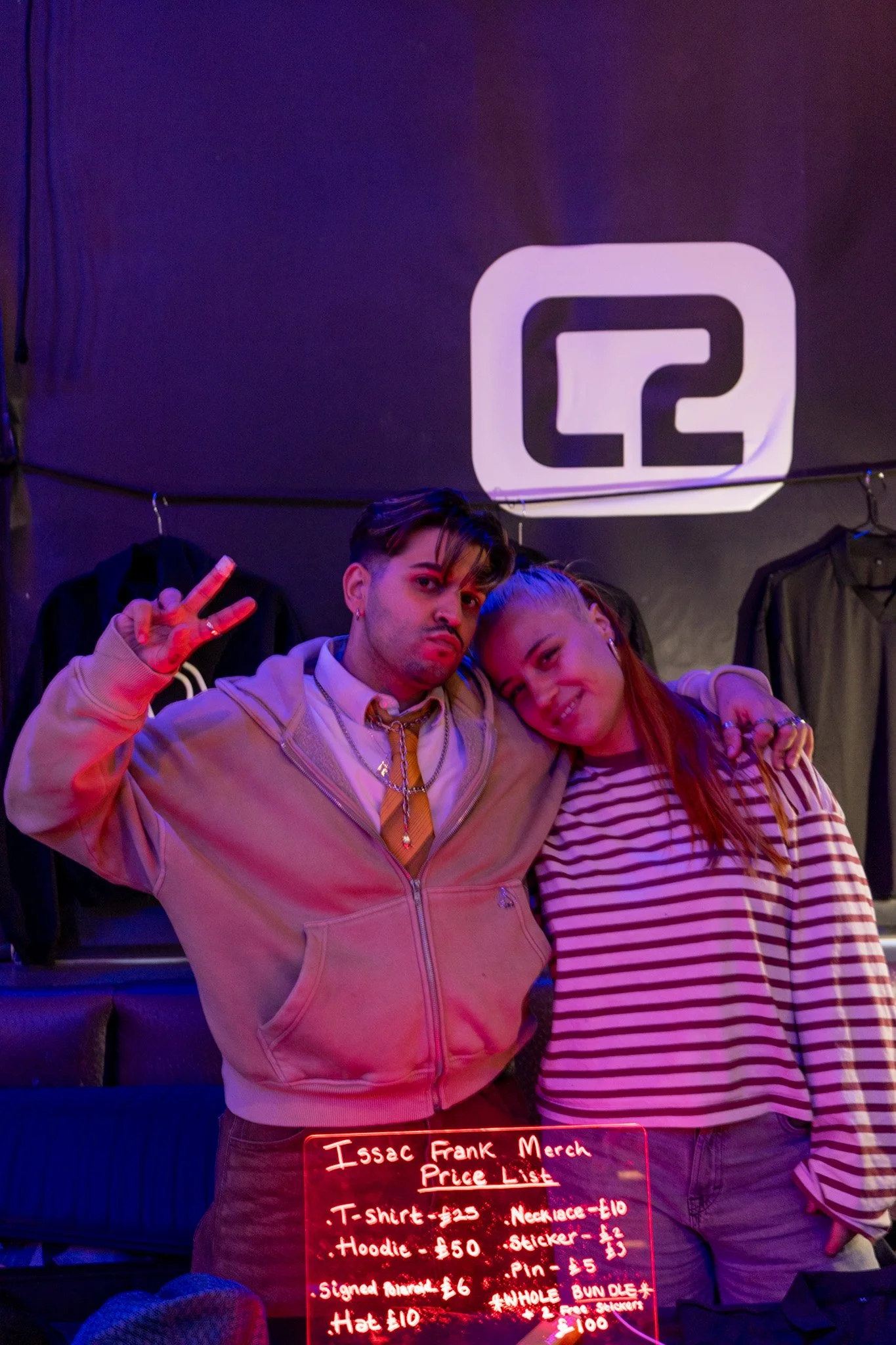 Two young adults, one male and one female, are standing close together in a dimly lit room with purple and red lighting. The male is making a peace sign with his right hand, wearing a beige hoodie, gold chain, and earrings. The female is leaning her 
