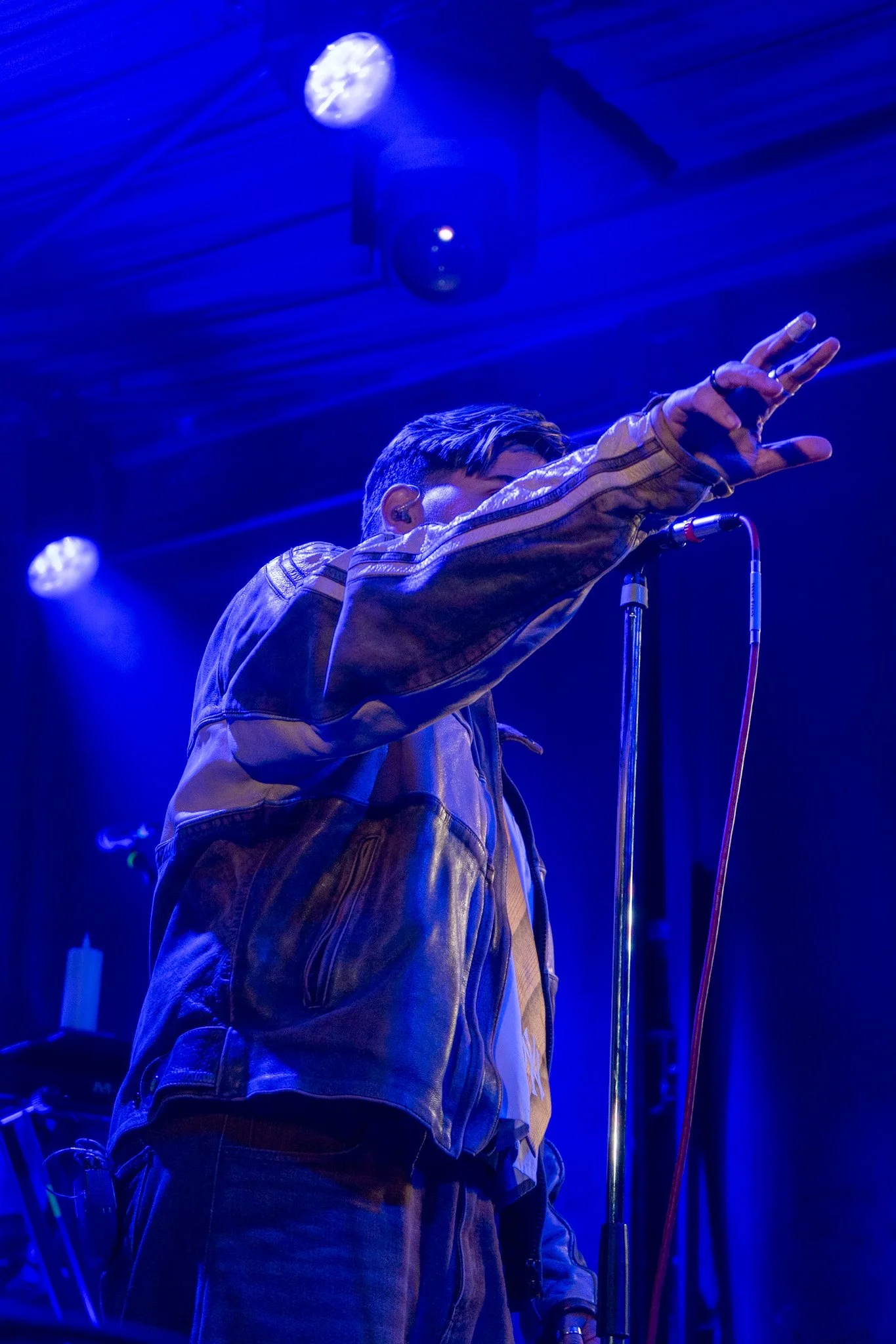 A person with short hair wearing a brown leather jacket and a white shirt, reaching towards the microphone stand in a dimly lit venue with blue lighting, stage lights, and equipment in the background.