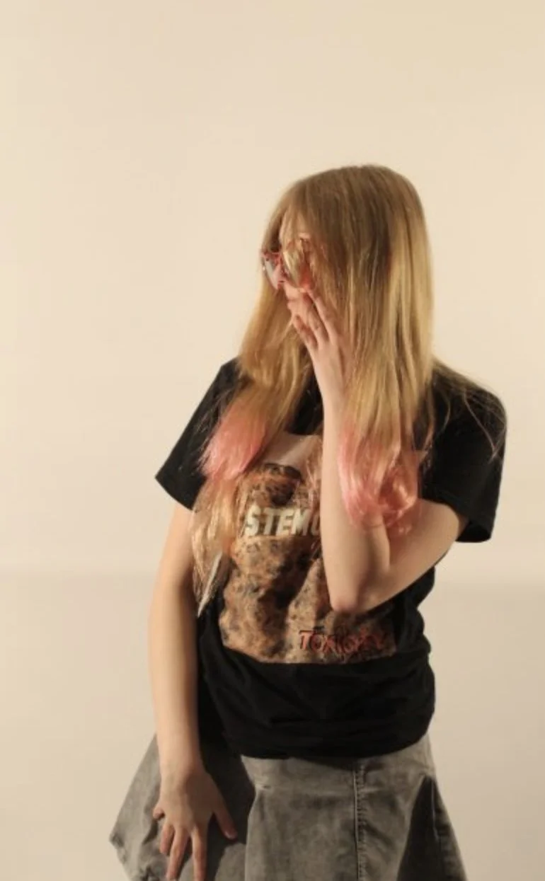 A young woman with long, blonde hair with pink tips, wearing glasses, a graphic t-shirt, and gray pants, covering her face with her hand against a plain, light-colored background.