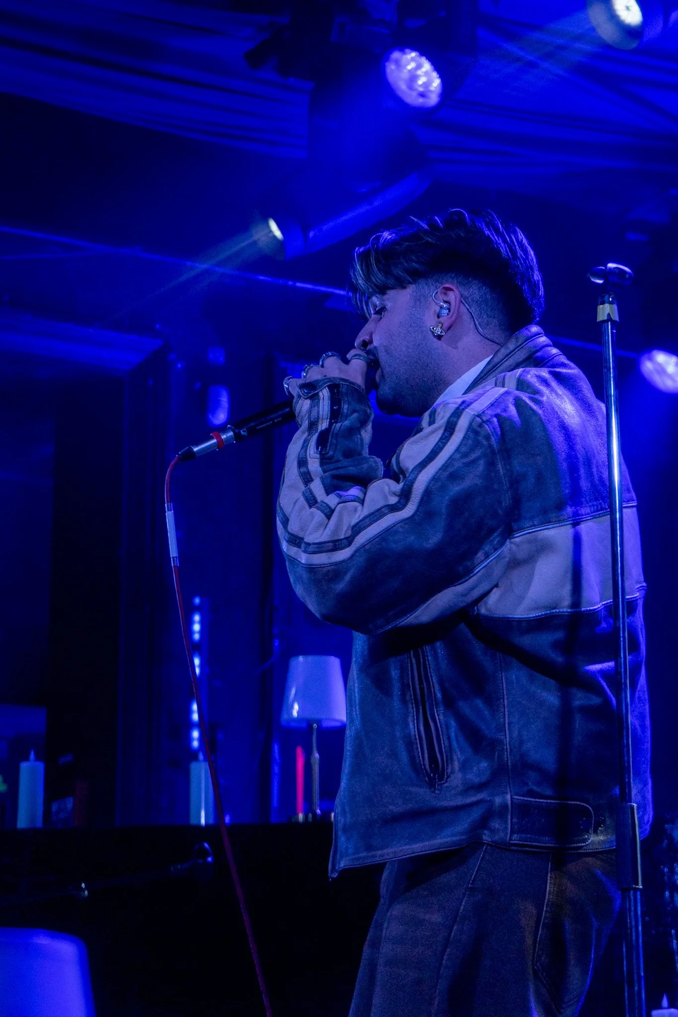 A male singer performing on stage under blue lighting. He is holding a microphone close to his mouth, wearing a denim jacket with striped sleeves and earrings, with a microphone stand beside him.