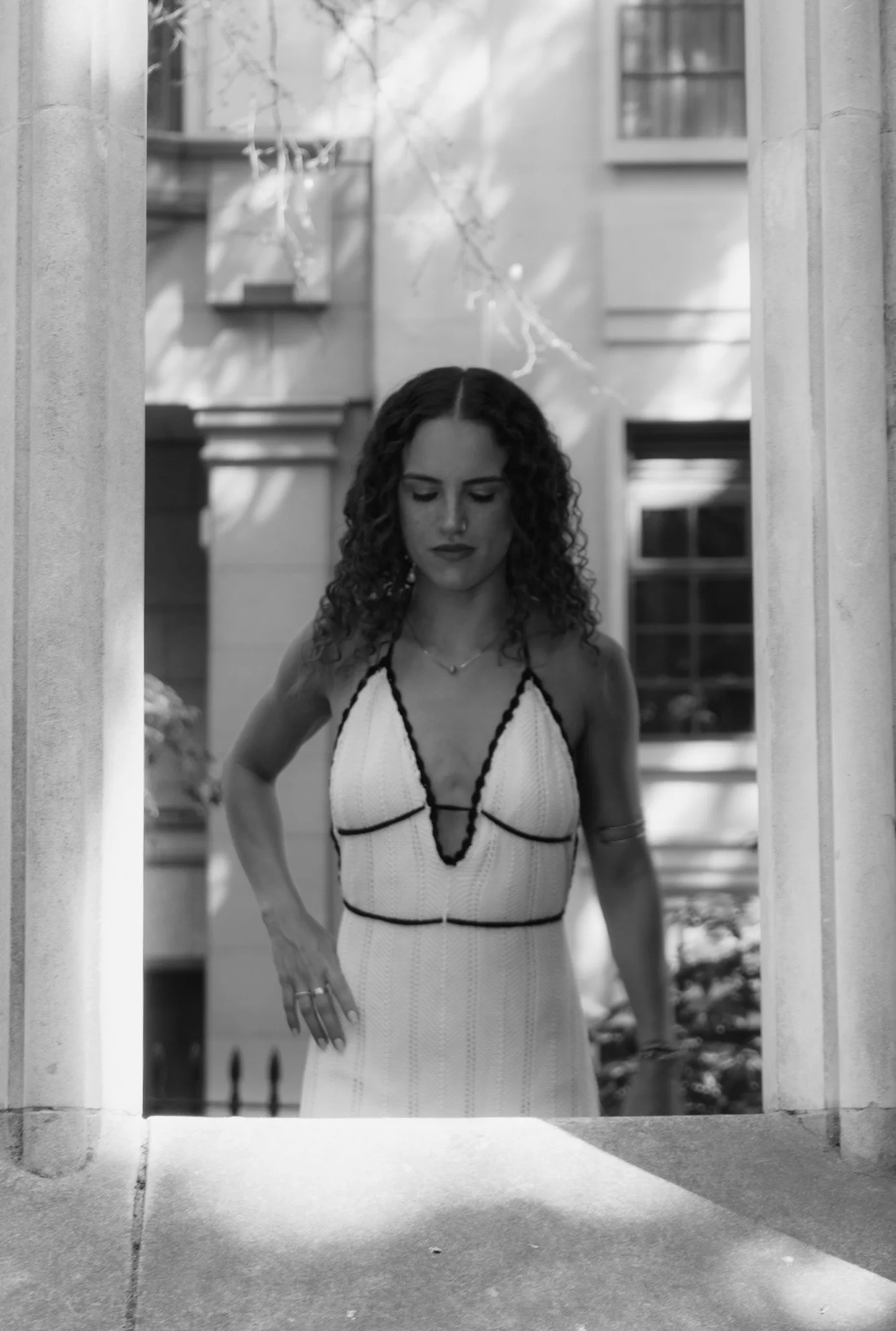 Black and white photo of a woman with curly hair wearing a patterned dress, looking downward, standing in an opening between two columns, with a house or building in the background.