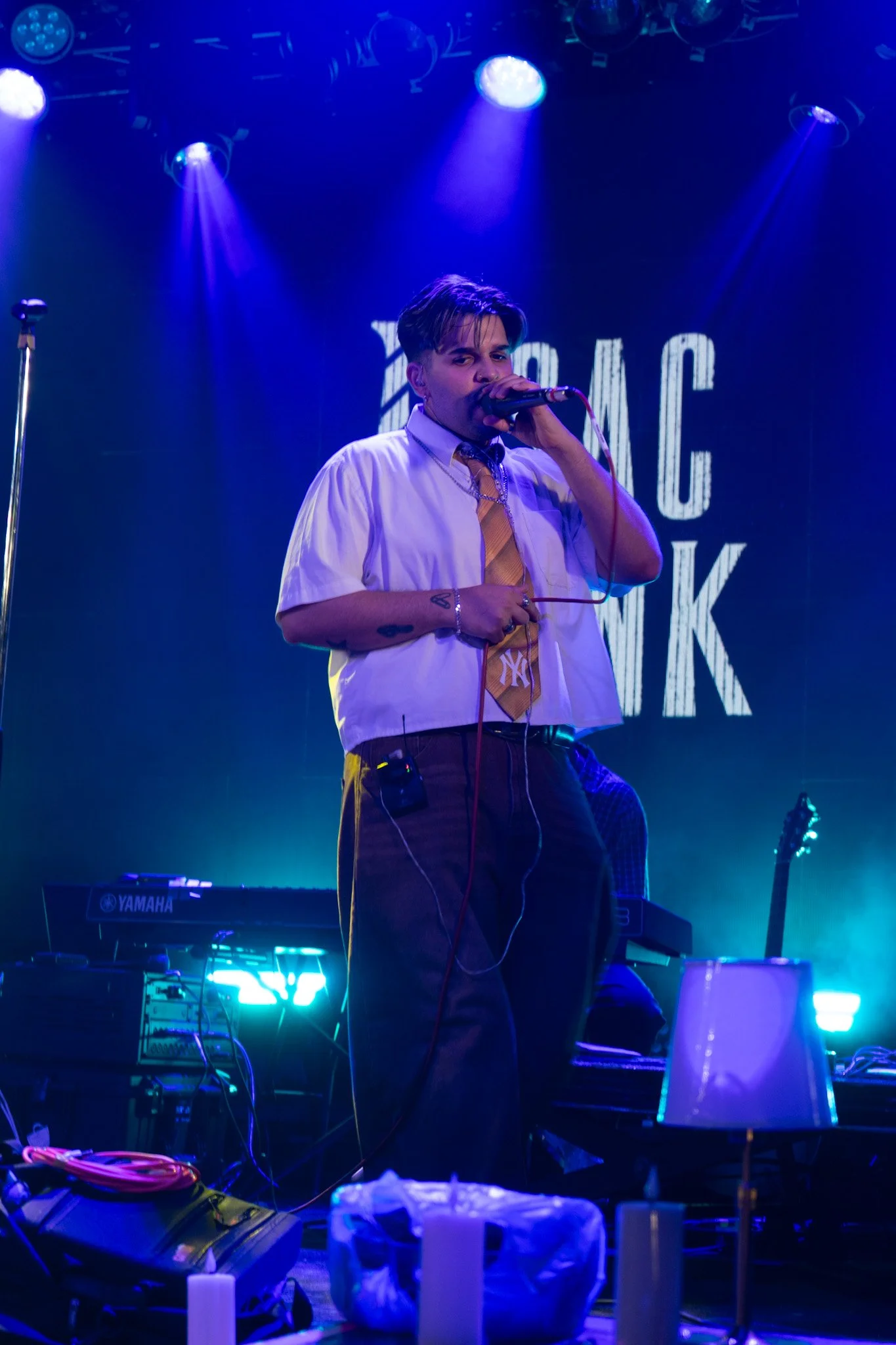 A male performer singing into a microphone on stage under purple and blue stage lights, with musical equipment and instruments visible in the background.