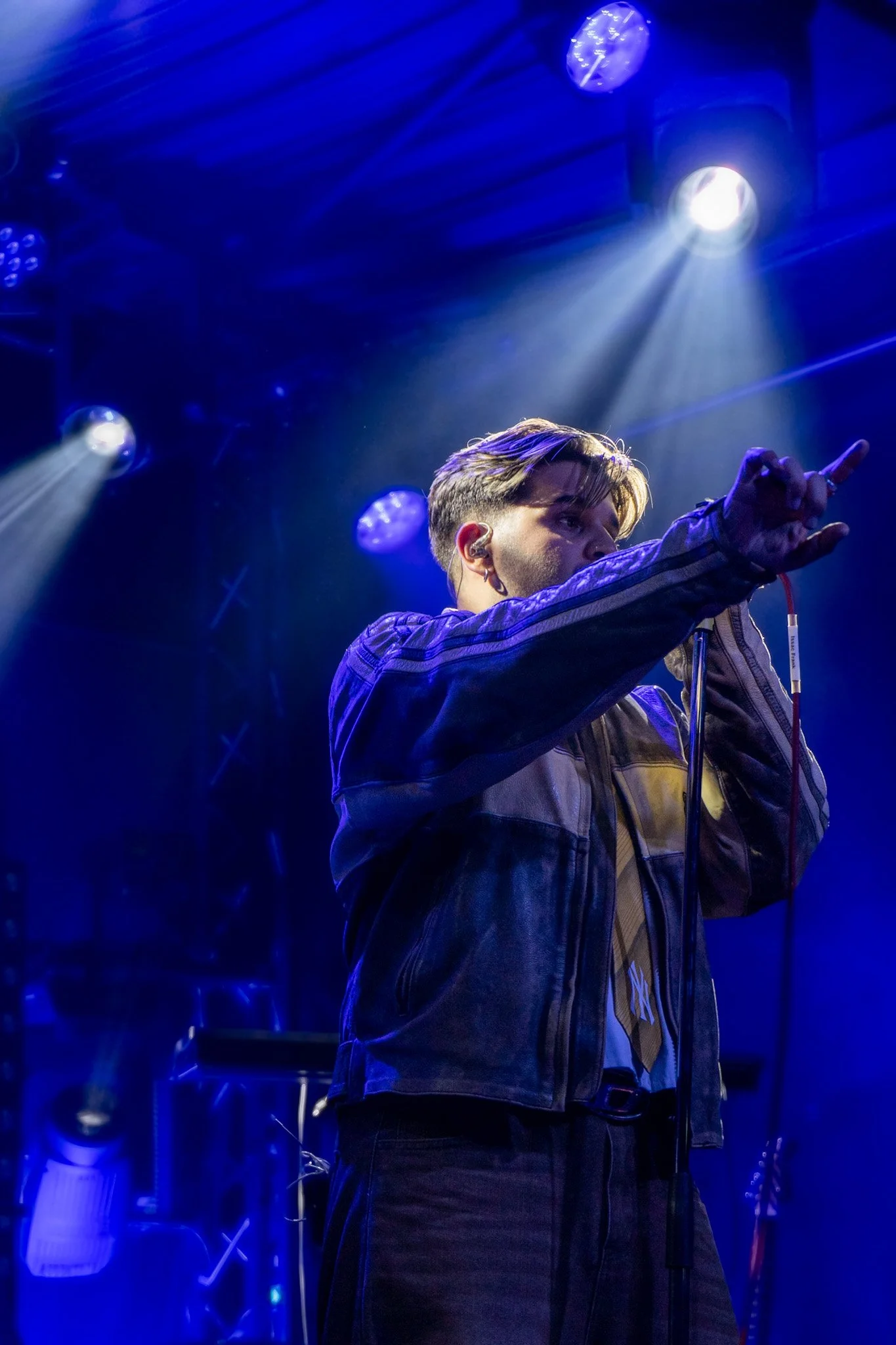 A young man with styled hair and a leather jacket performing on stage under blue and white stage lights, with a clock and various equipment in the background.