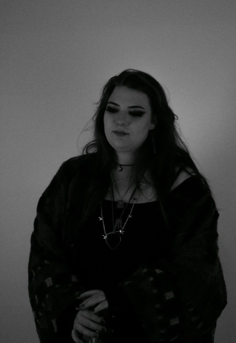 A young woman with dark hair and makeup, wearing layered necklaces, a dark top, and a plaid jacket, standing against a plain background, with arms crossed.