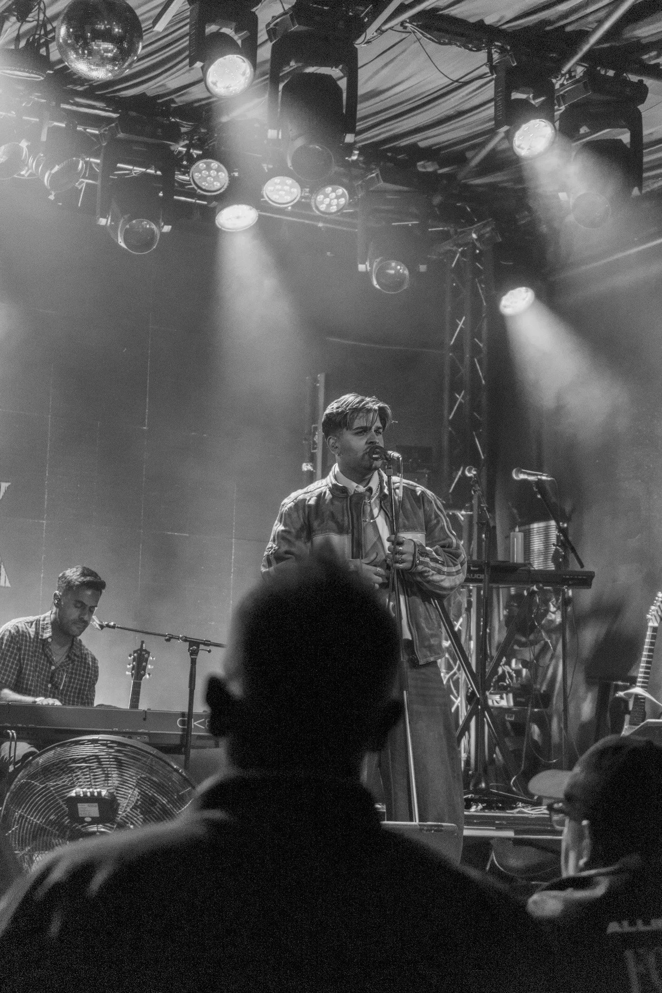 A black and white photo of a live music performance with a singer at the microphone, a keyboard player, and various stage equipment, with spotlights and a disco ball hanging from the ceiling.