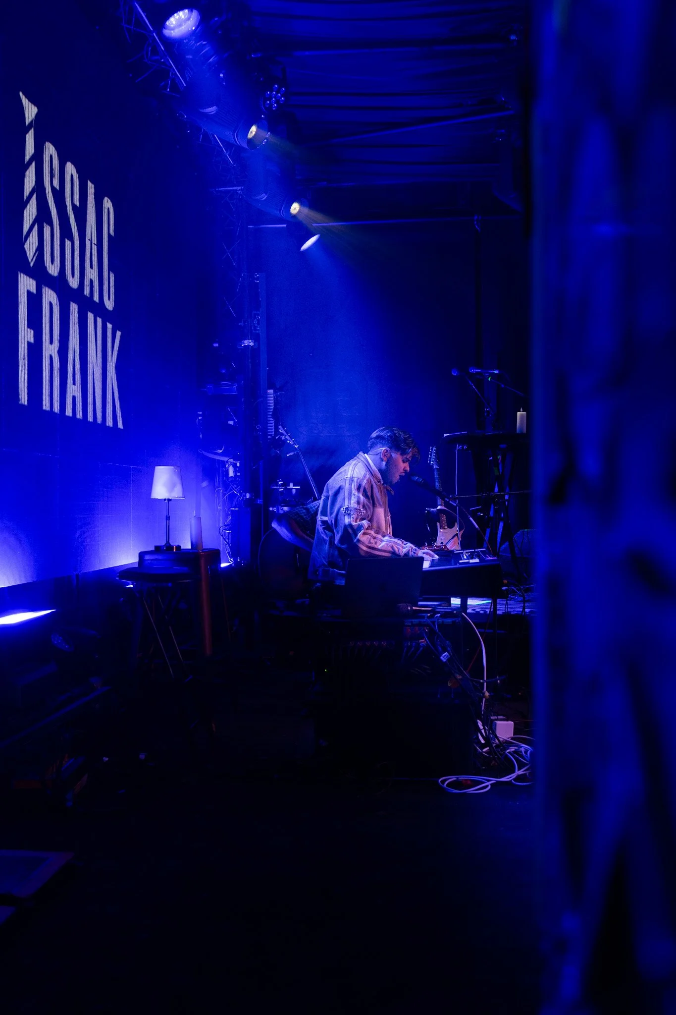 A musician performing on a stage illuminated with blue lighting, with a large screen behind displaying the name 'HUSH FRANK'. The stage has various musical equipment, and the musician appears to be singing into a microphone while playing an instrumen