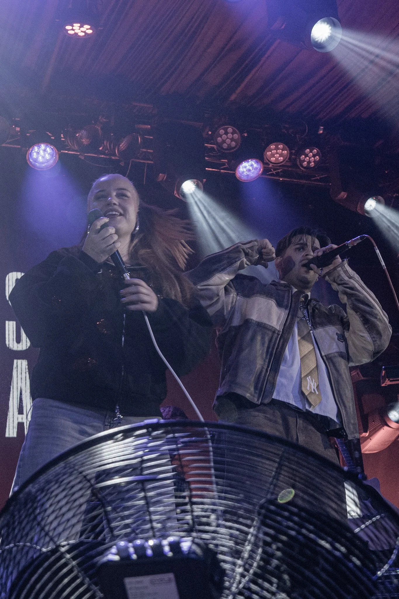 Two performers on stage with microphones, purple and blue stage lights, a large fan in the foreground, and a dark backdrop.