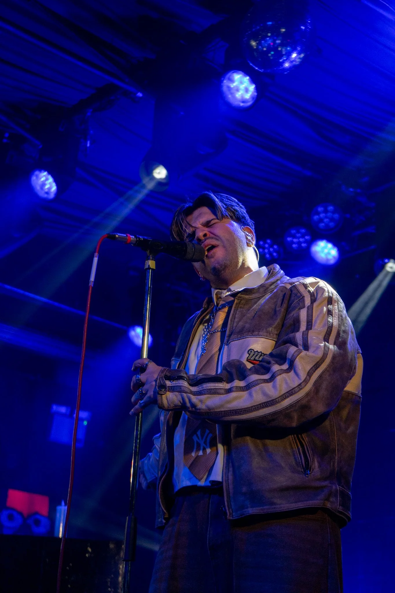 Male singer performing on stage under blue lights, with a microphone and music equipment in the background.