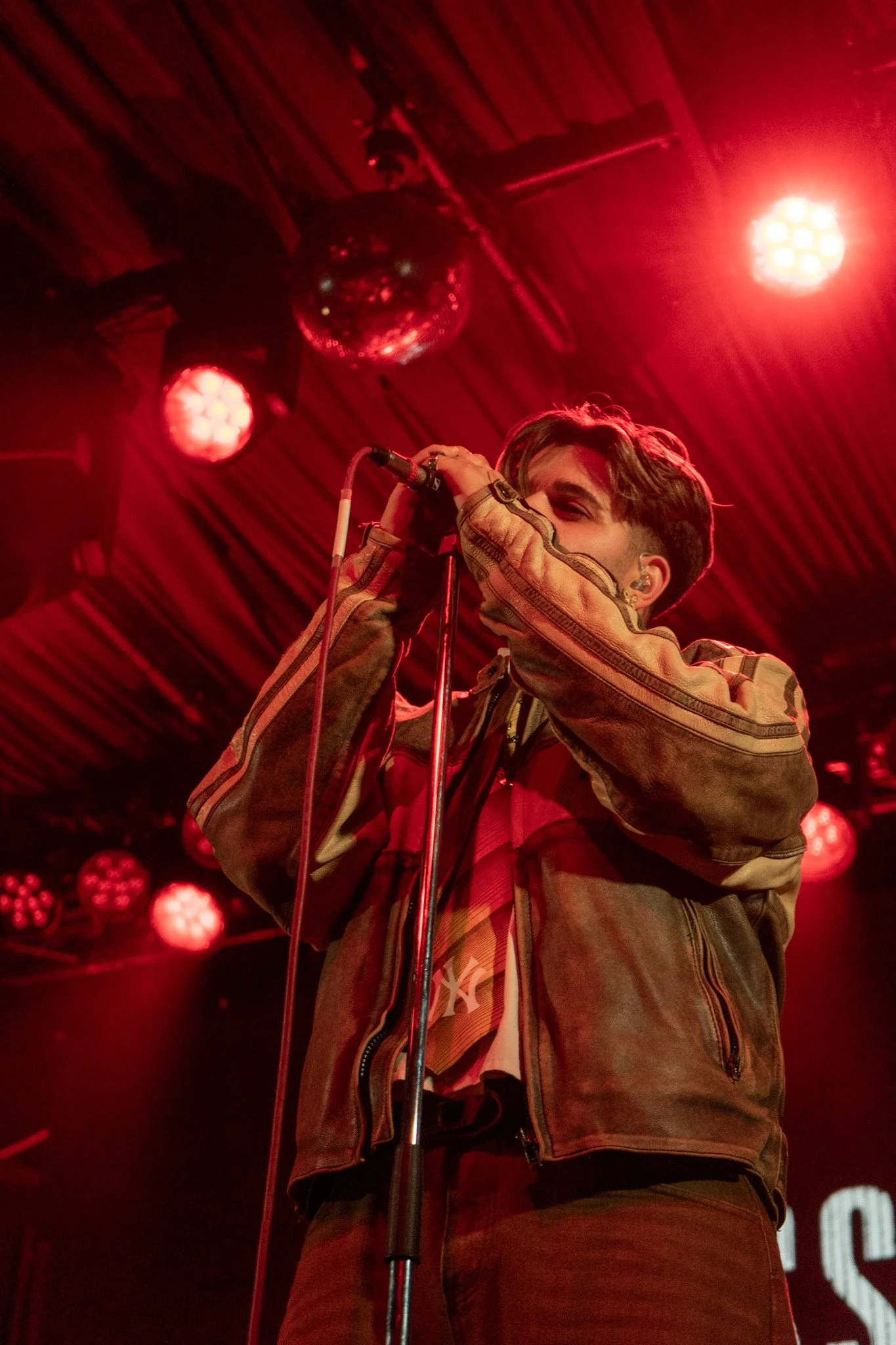 A person with brown hair, wearing a brown jacket, singing into a microphone on stage with red lighting and a disco ball hanging from the ceiling.
