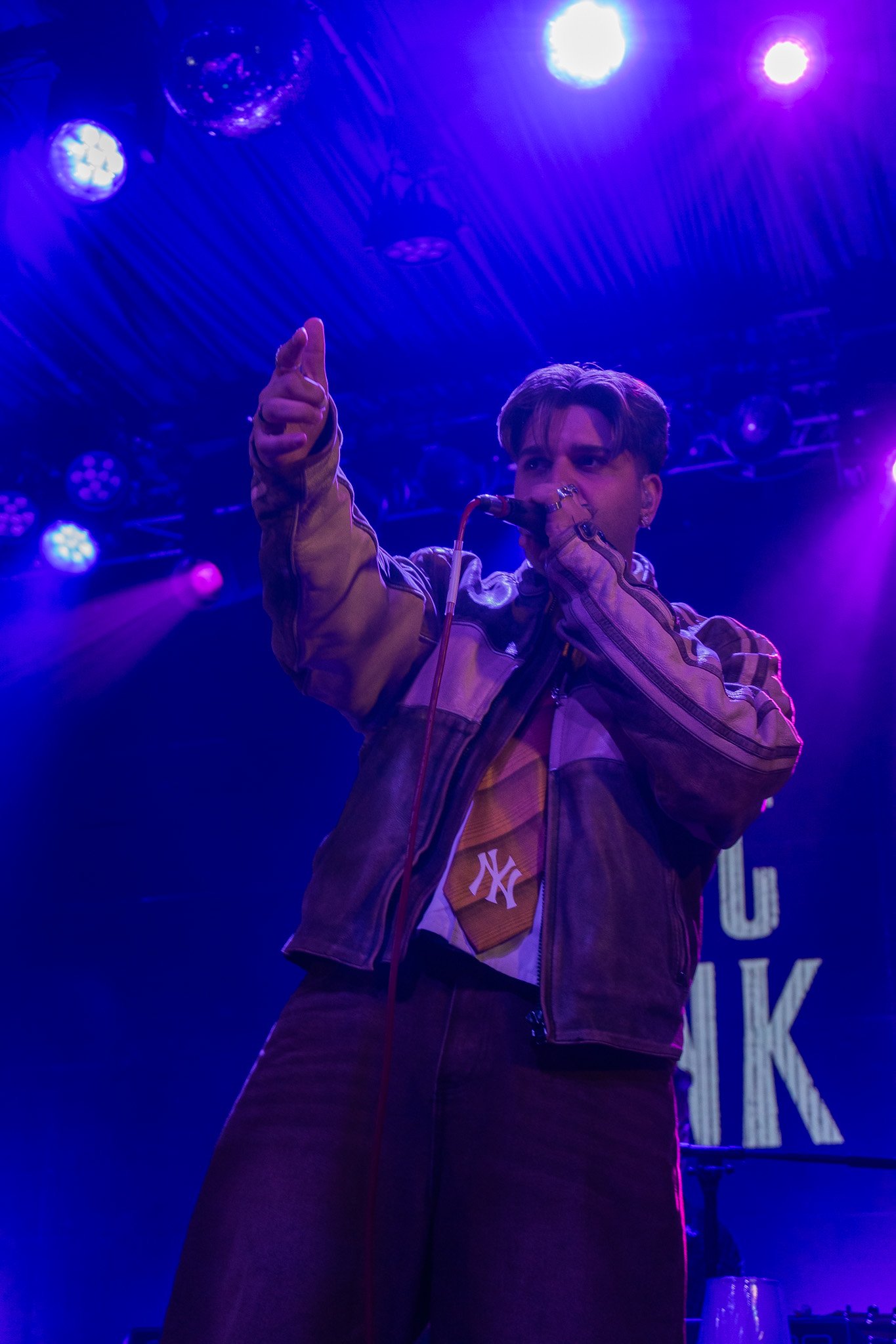 A male singer performing on stage, wearing a brown jacket with a NY logo and pointing towards the camera, with purple and blue stage lights overhead.
