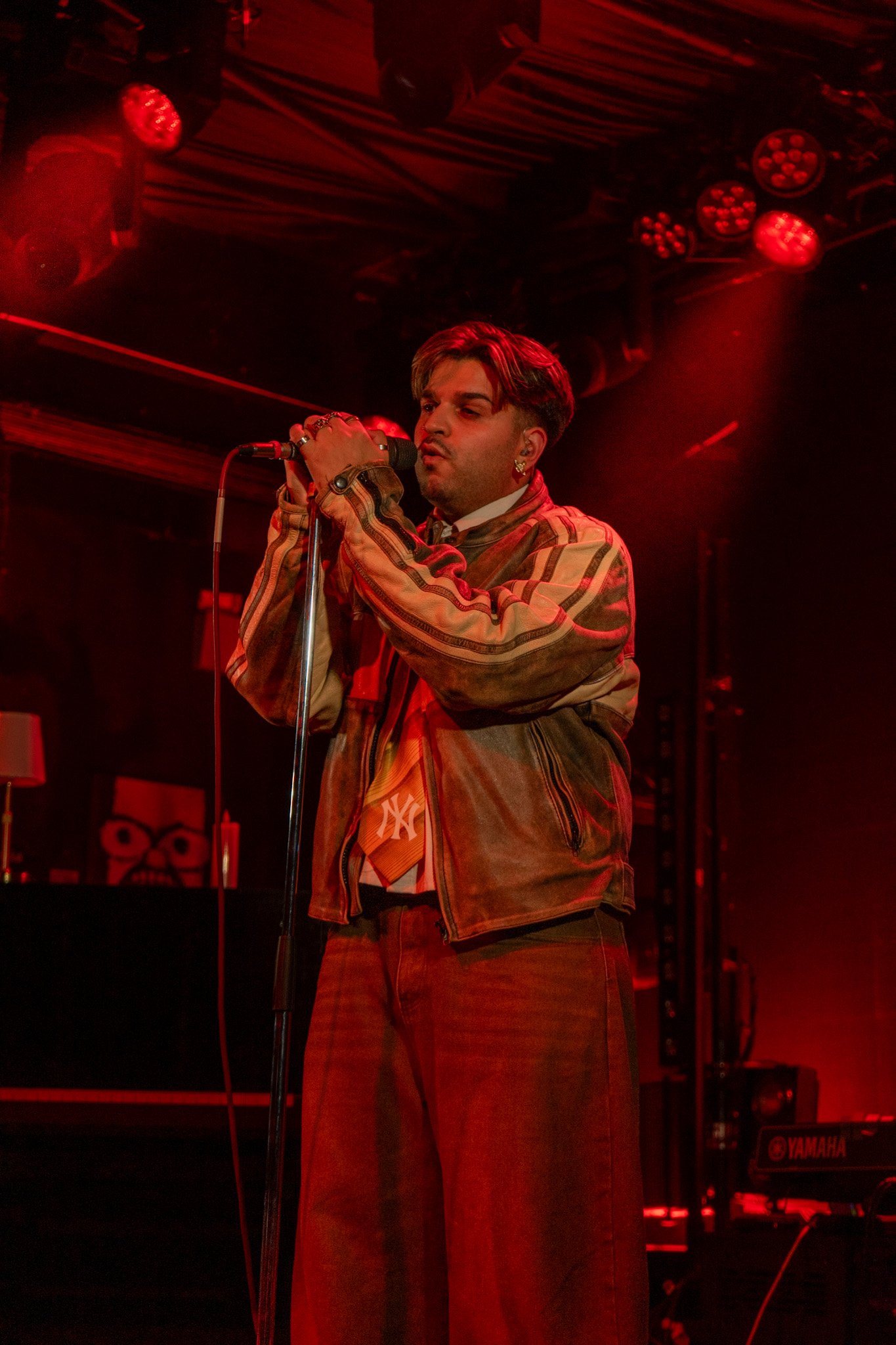 Male singer performing on stage under red lighting, wearing a plaid jacket and brown pants with a necktie, holding a microphone.