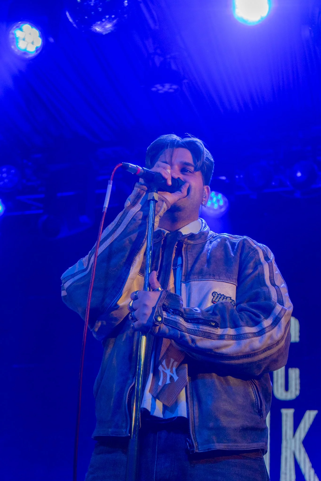 A male performer singing into a microphone on stage, illuminated by blue stage lights, wearing a brown leather jacket.