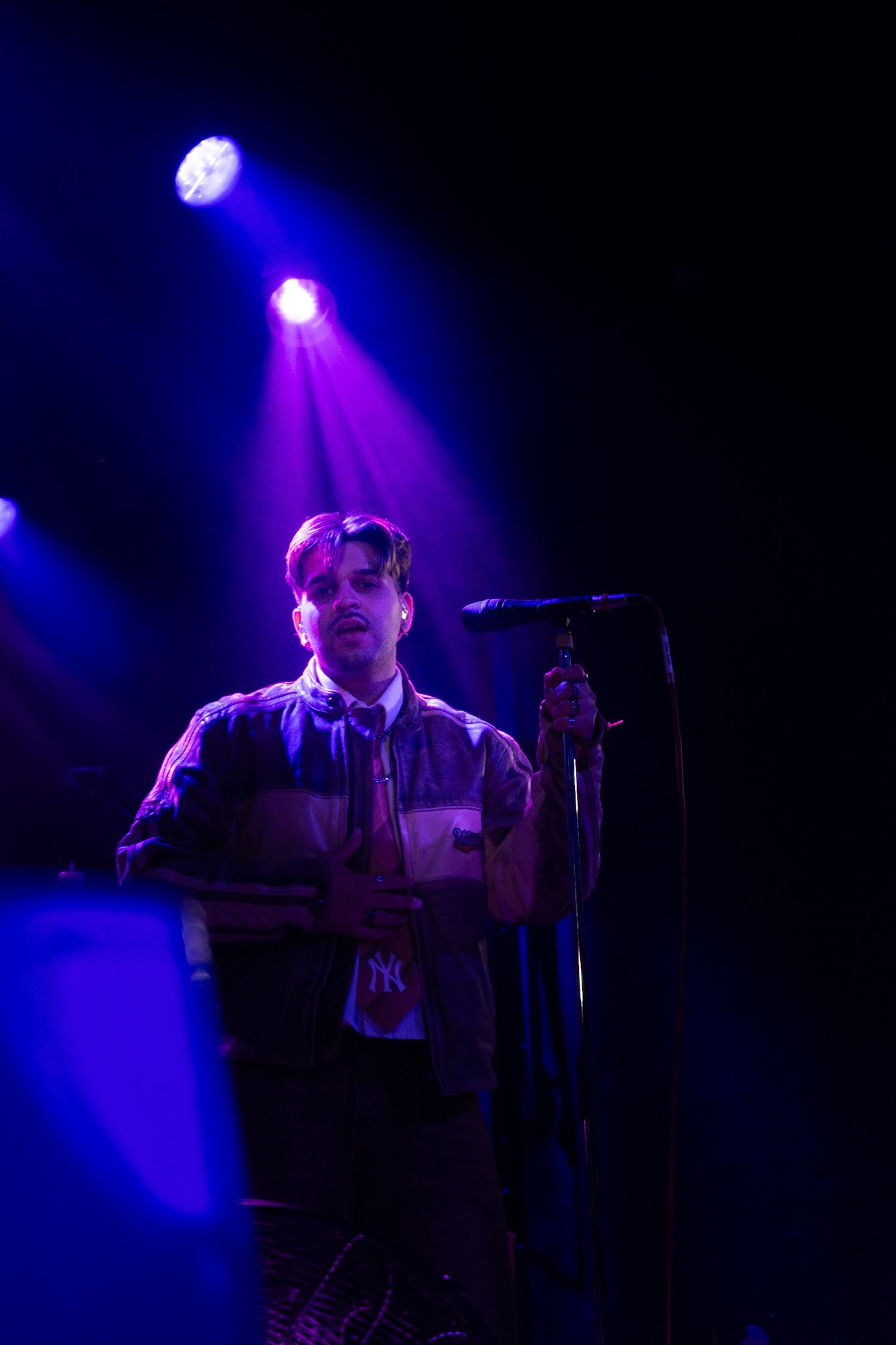A male singer performing on stage under blue and purple stage lights, holding a microphone in his right hand. He is wearing a striped jacket with a collar and a shirt underneath. The background is dark with stage equipment visible.