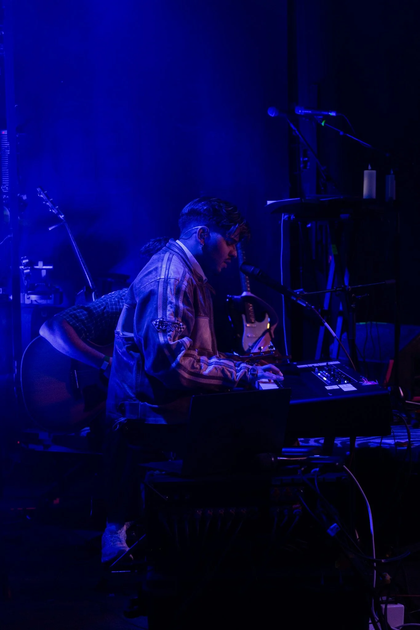A man with glasses sitting at a keyboard on a stage, illuminated by blue stage lighting, with microphones and music equipment around him.