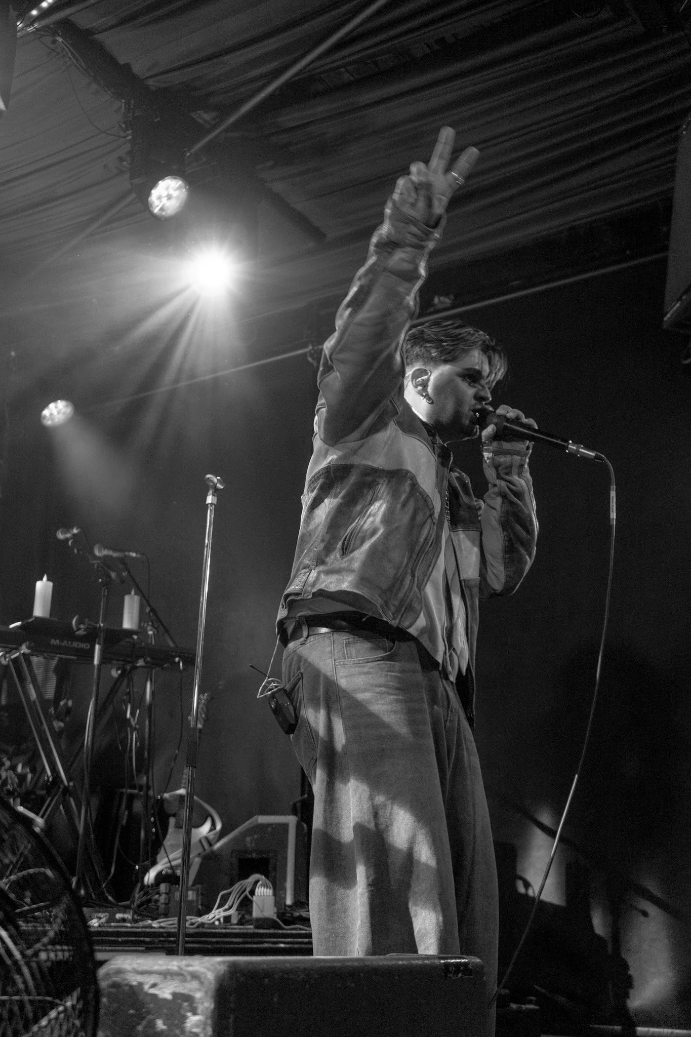 A male singer on stage holding a microphone and making a peace sign with his hand. He is wearing a jacket and standing near music equipment and microphones in a dimly lit venue.