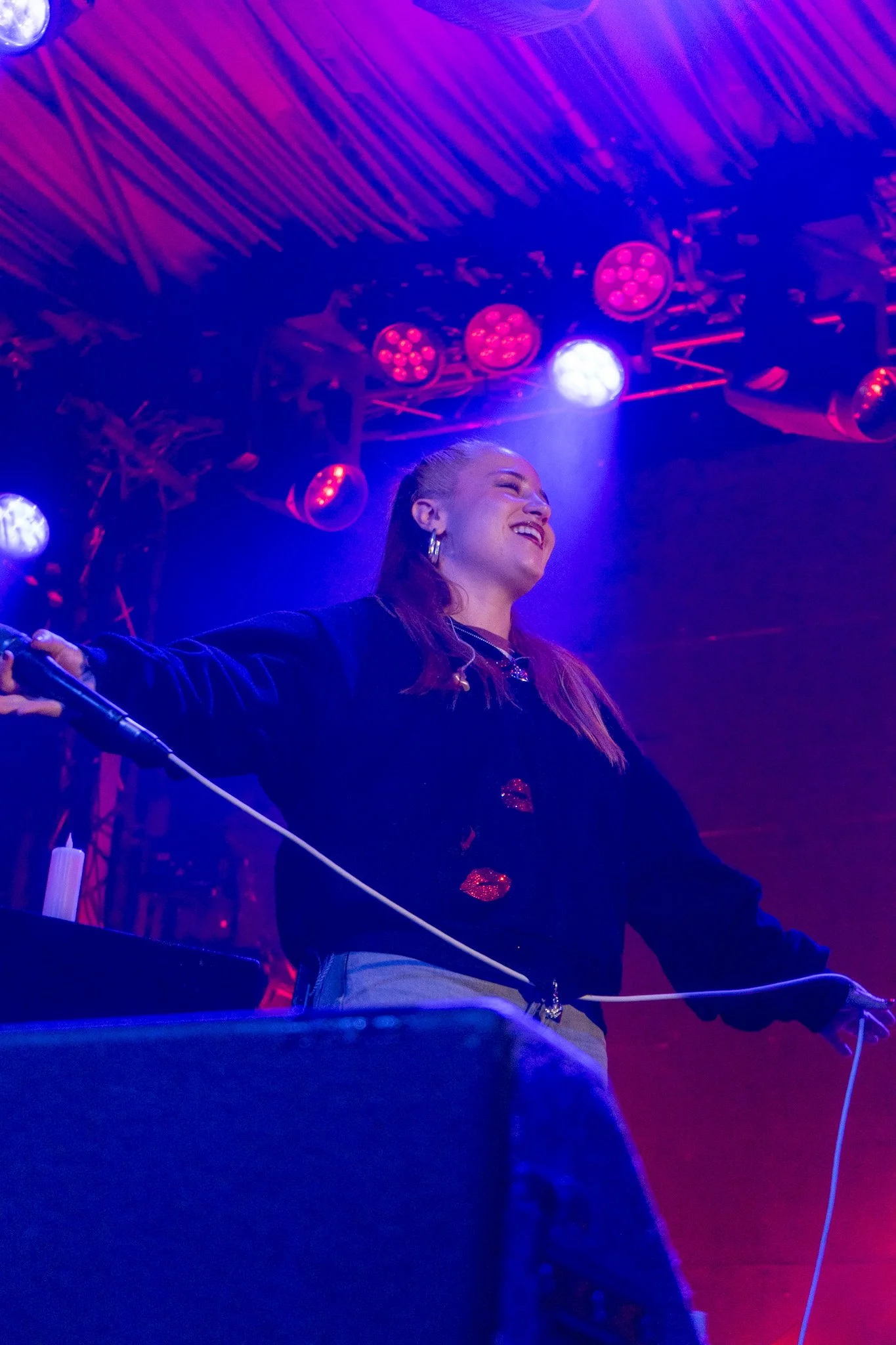 Person with long hair smiling under colorful stage lights, wearing a dark jacket with red lips pattern, on stage with equipment and lighting rig.
