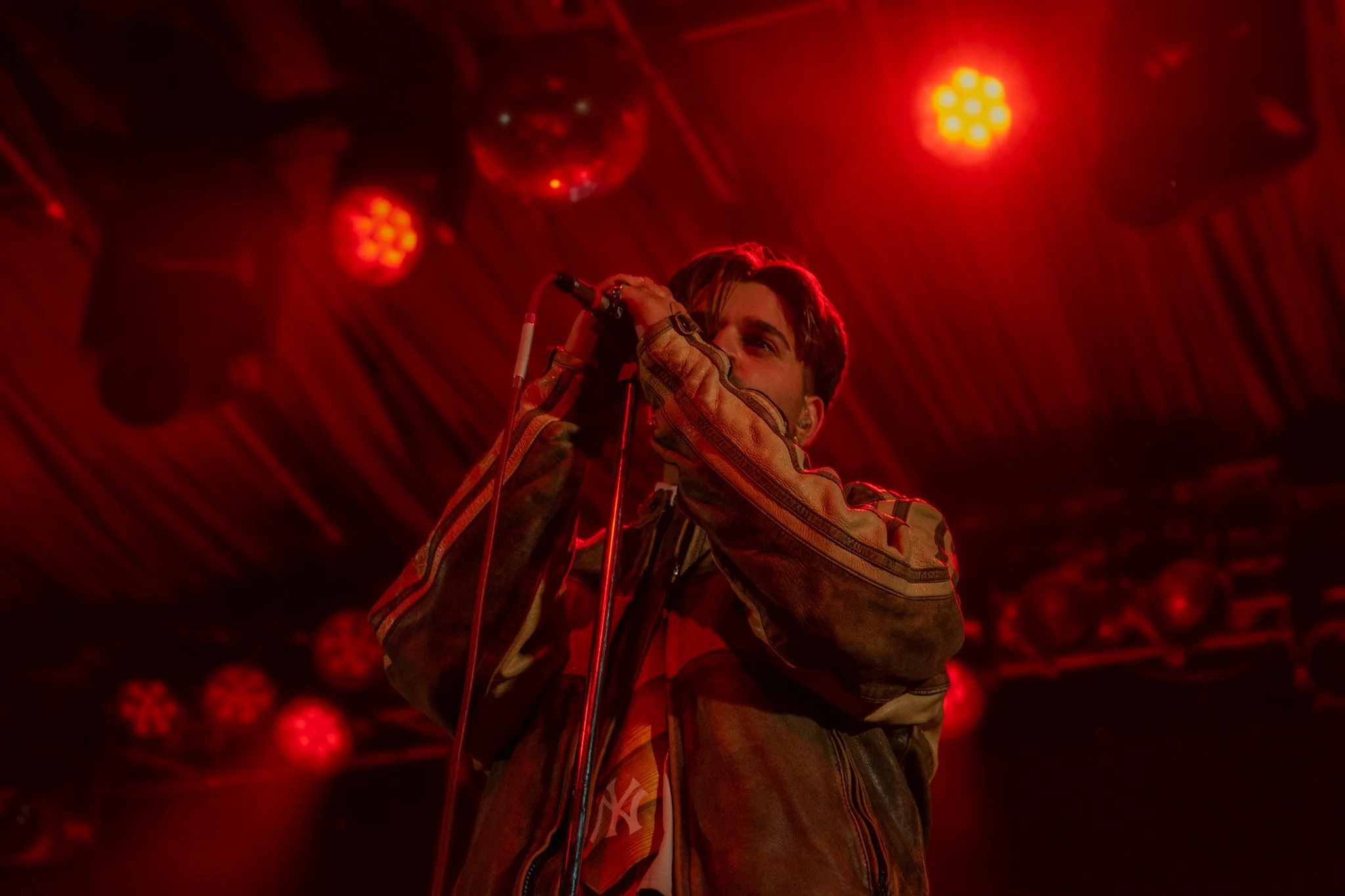 A male performer singing into a microphone on stage illuminated by red lights.