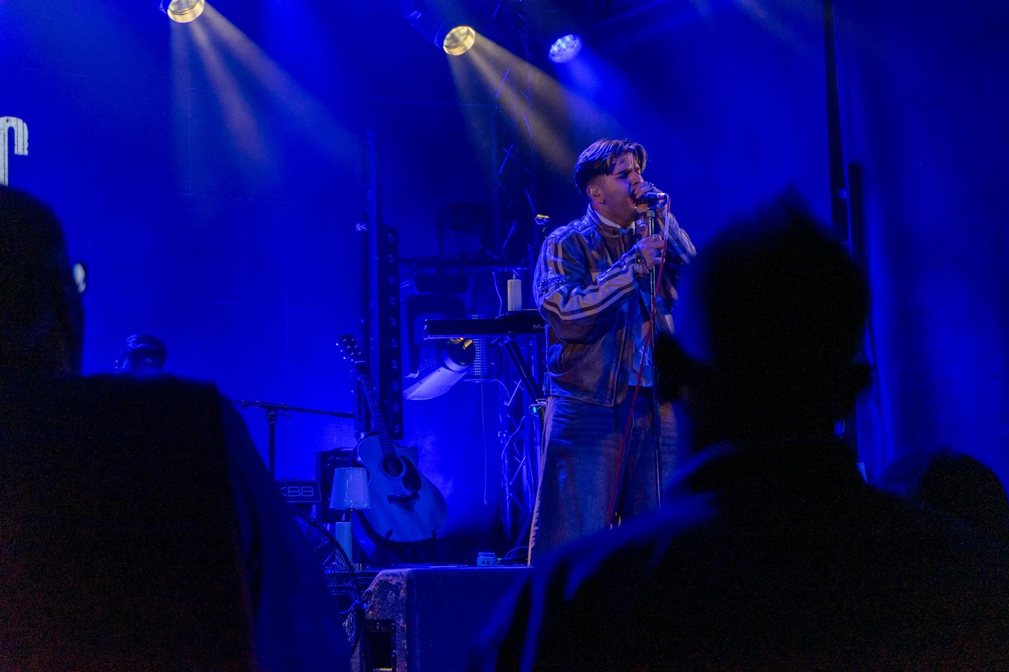 A male singer with short hair and earrings, wearing a striped jacket, performs on stage with blue lighting. There is musical equipment, including a keyboard and guitars, visible behind him.