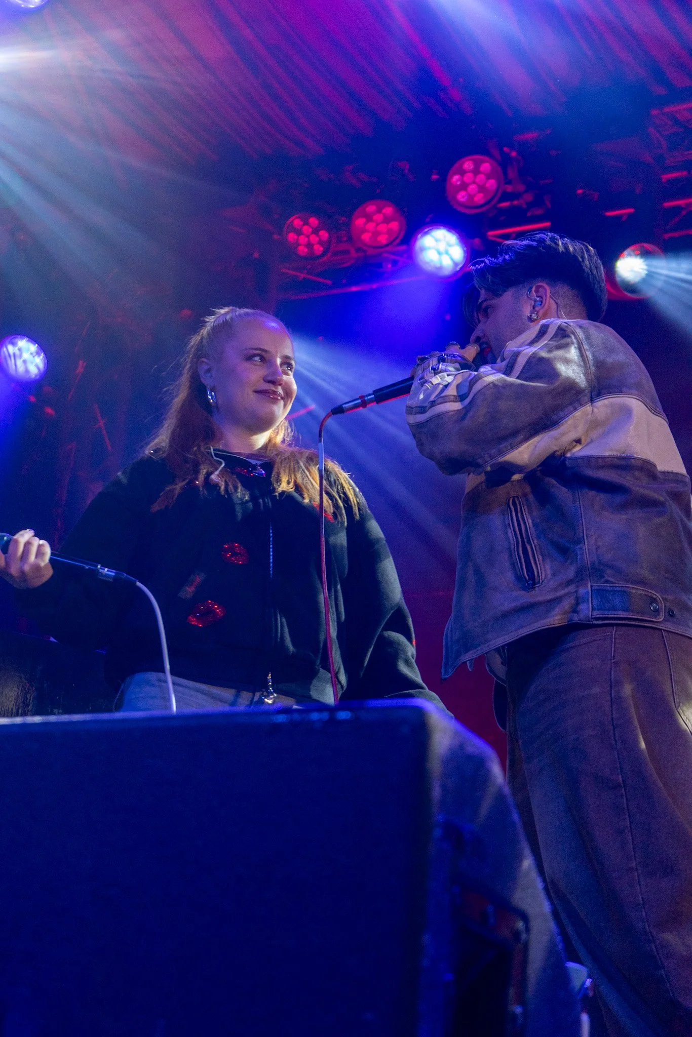 Two musicians perform on stage under colored lights, one holding a microphone and the other with a face partially visible, in a lively concert setting.