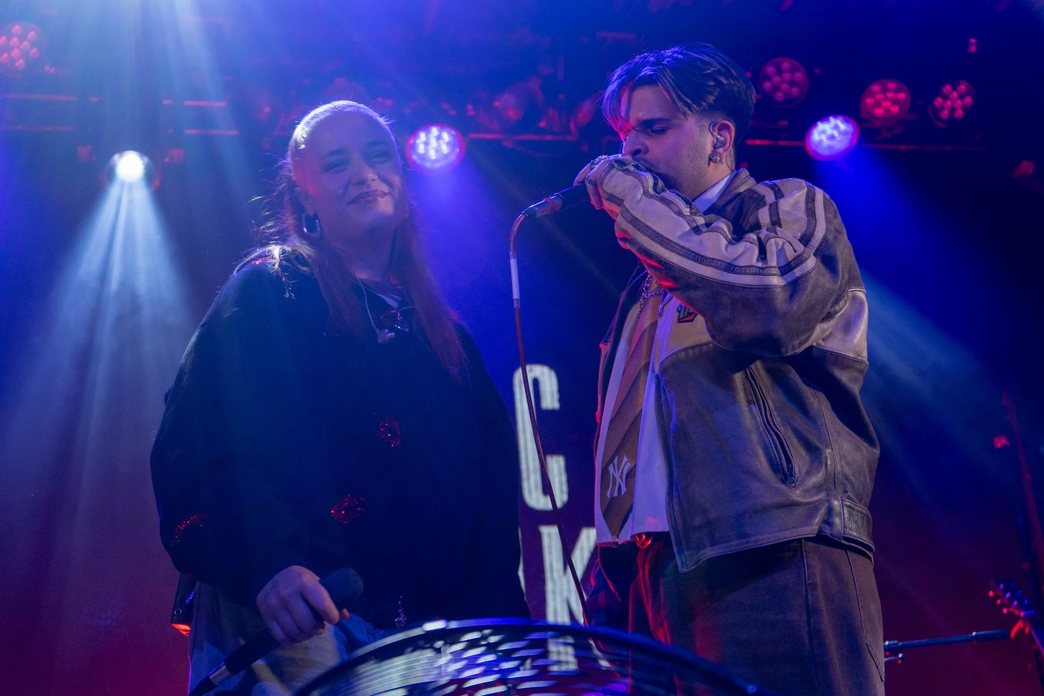 A man singing into a microphone on stage while a woman stands nearby with a smile, under colorful stage lights.