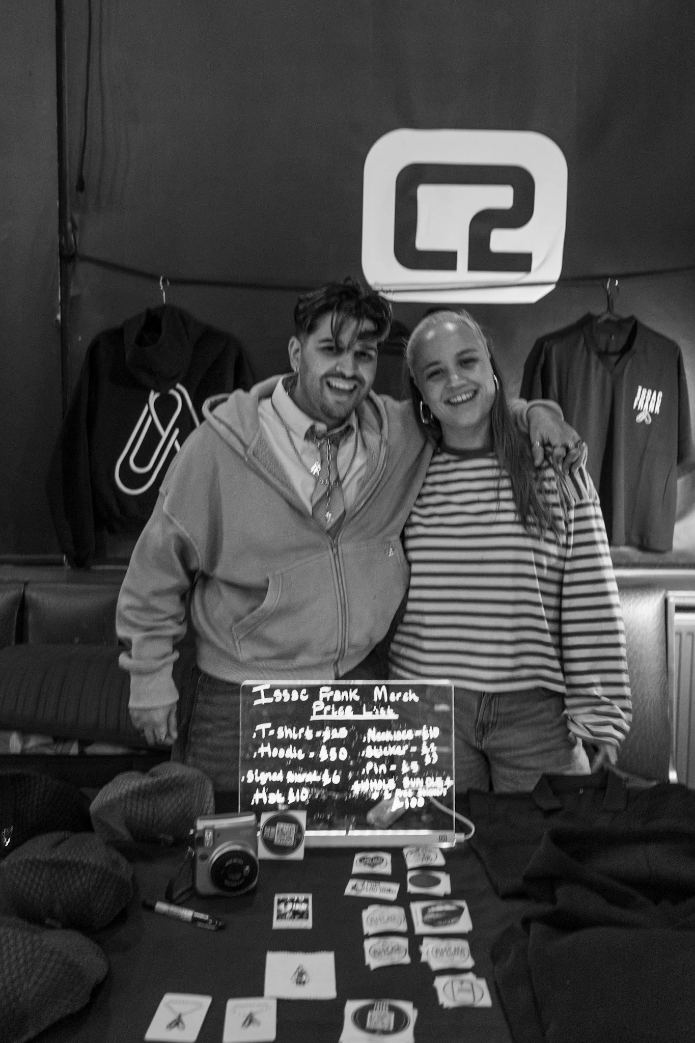 Two people smiling, standing behind a table with hand-drawn signs and cards, at an indoor event with a sign that reads 'Q2' in the background.