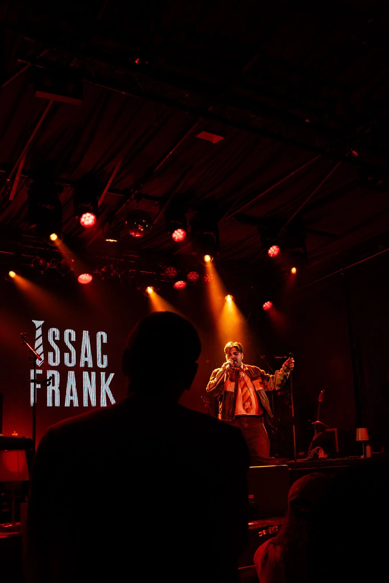 A singer performing on stage with red and orange stage lights, with a sign that reads 'Issac Frank' in the background.