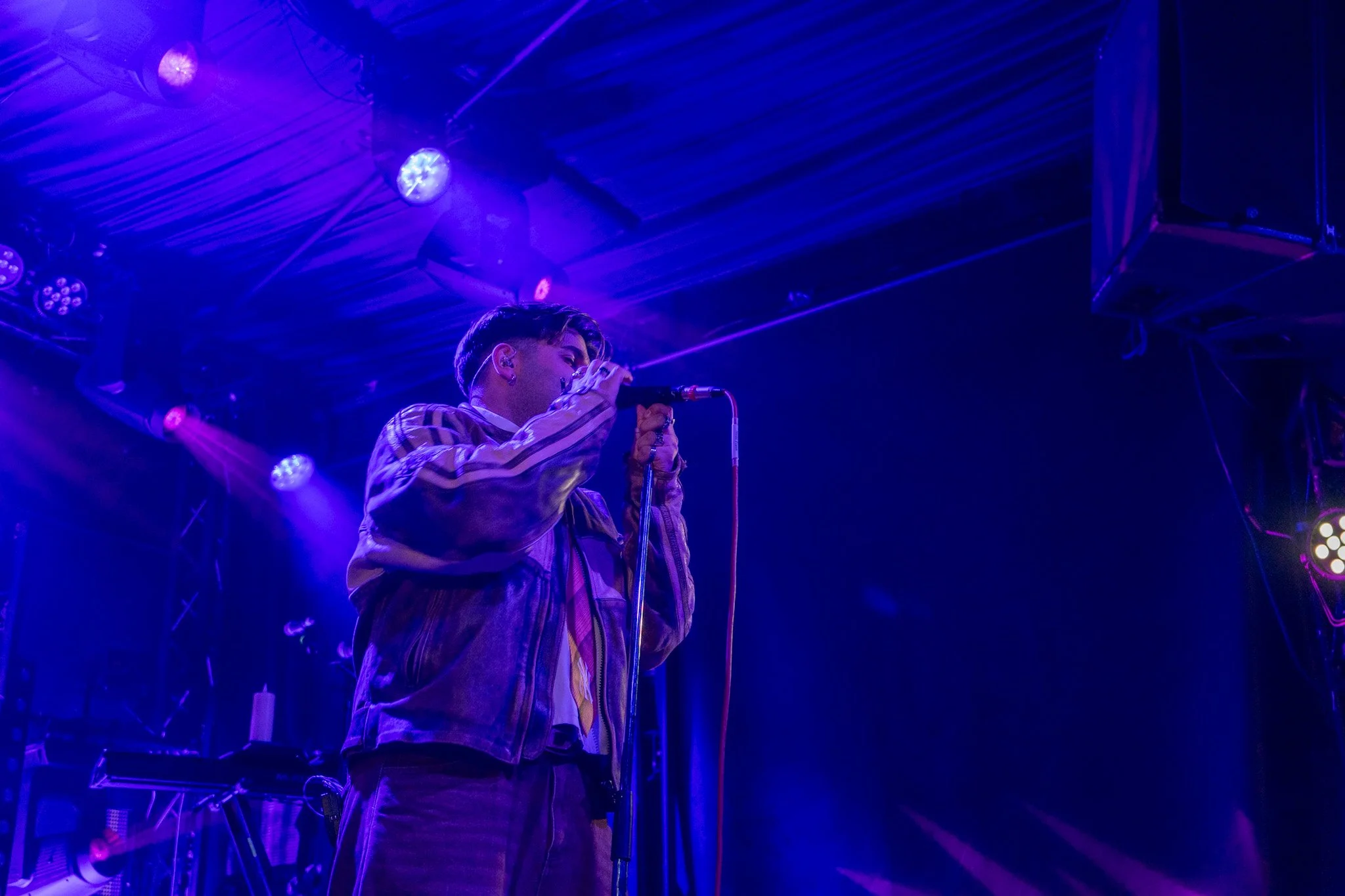 A person singing into a microphone on a stage illuminated with purple and blue lights.