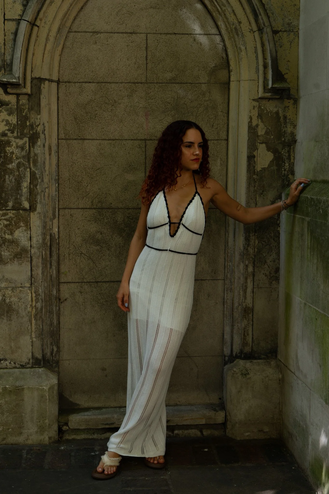 A woman with curly hair wearing a white dress with black accents, standing in front of a stone wall and arched doorway, touching the wall with her right hand.
