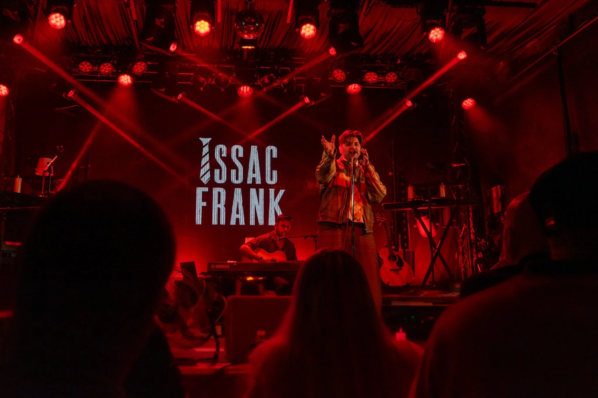 Live music performance with a singer and guitarist on stage with red lighting and a large screen displaying 'ISSAC FRANK' in the background.