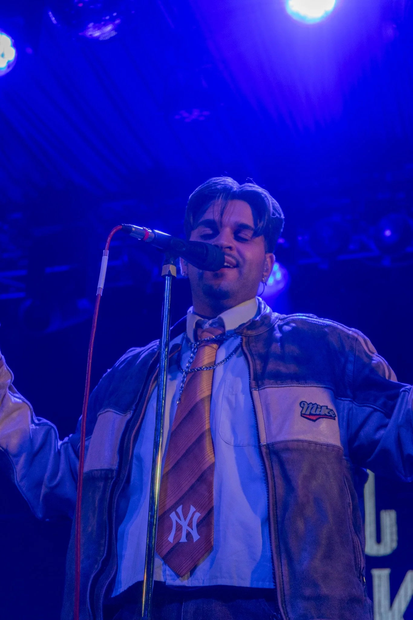 A man with short hair and earrings singing into a microphone on stage with blue lighting, wearing a varsity jacket with a NY logo and a NY Yankees shirt underneath.