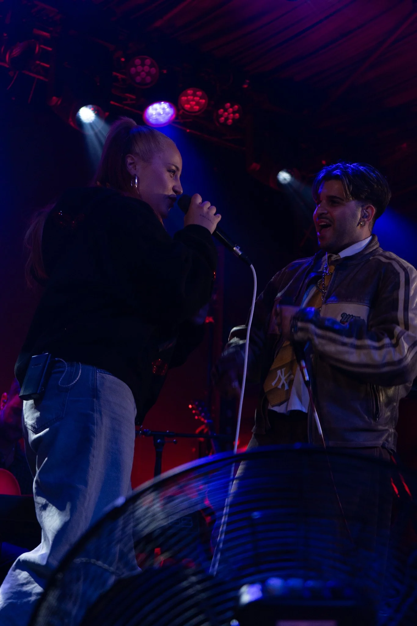 Two singers performing on stage with colorful lights above them, one woman holding a microphone and a man with earbuds, engaging with the audience.