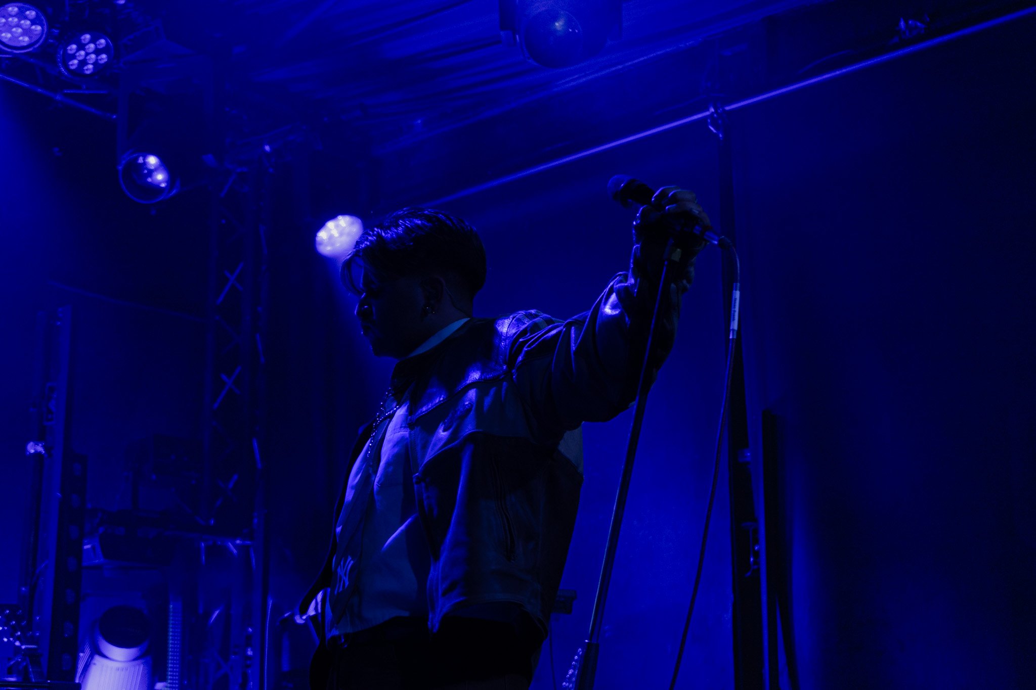 A person holding a microphone on stage with blue lighting, stage equipment and scaffolding visible in the background.