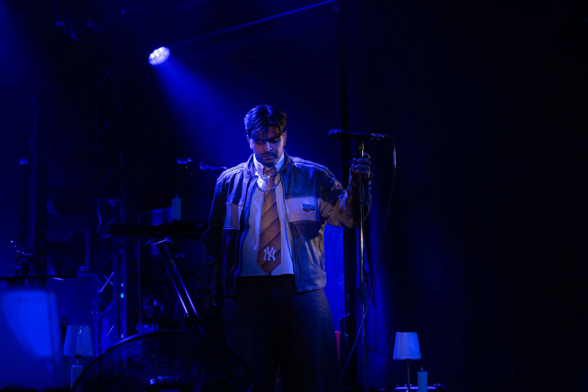A male singer performing on stage under blue lighting, wearing a jacket and holding a microphone stand.