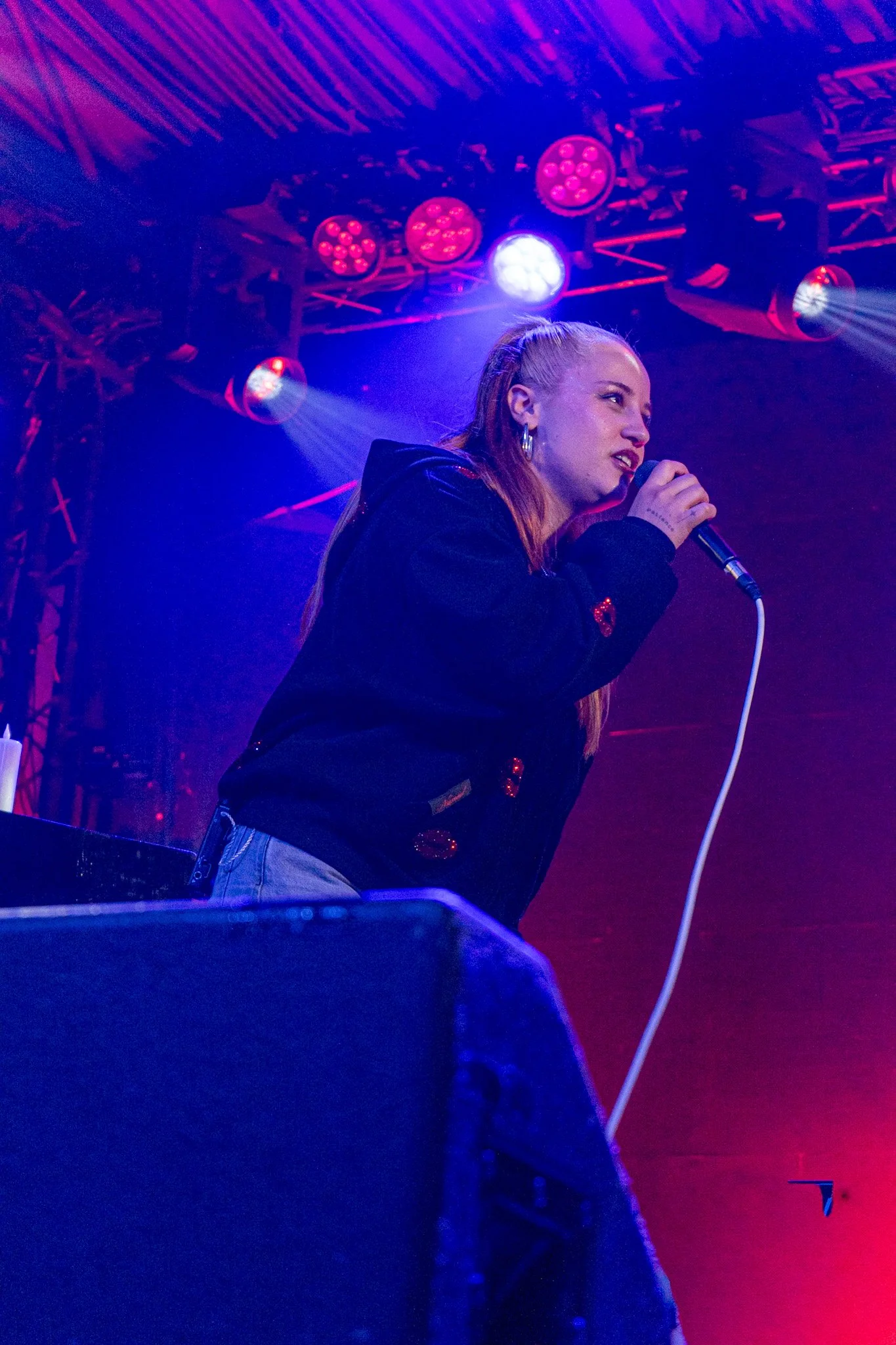 A female singer with red hair performing on stage under pink and blue stage lights, holding a microphone near her mouth.