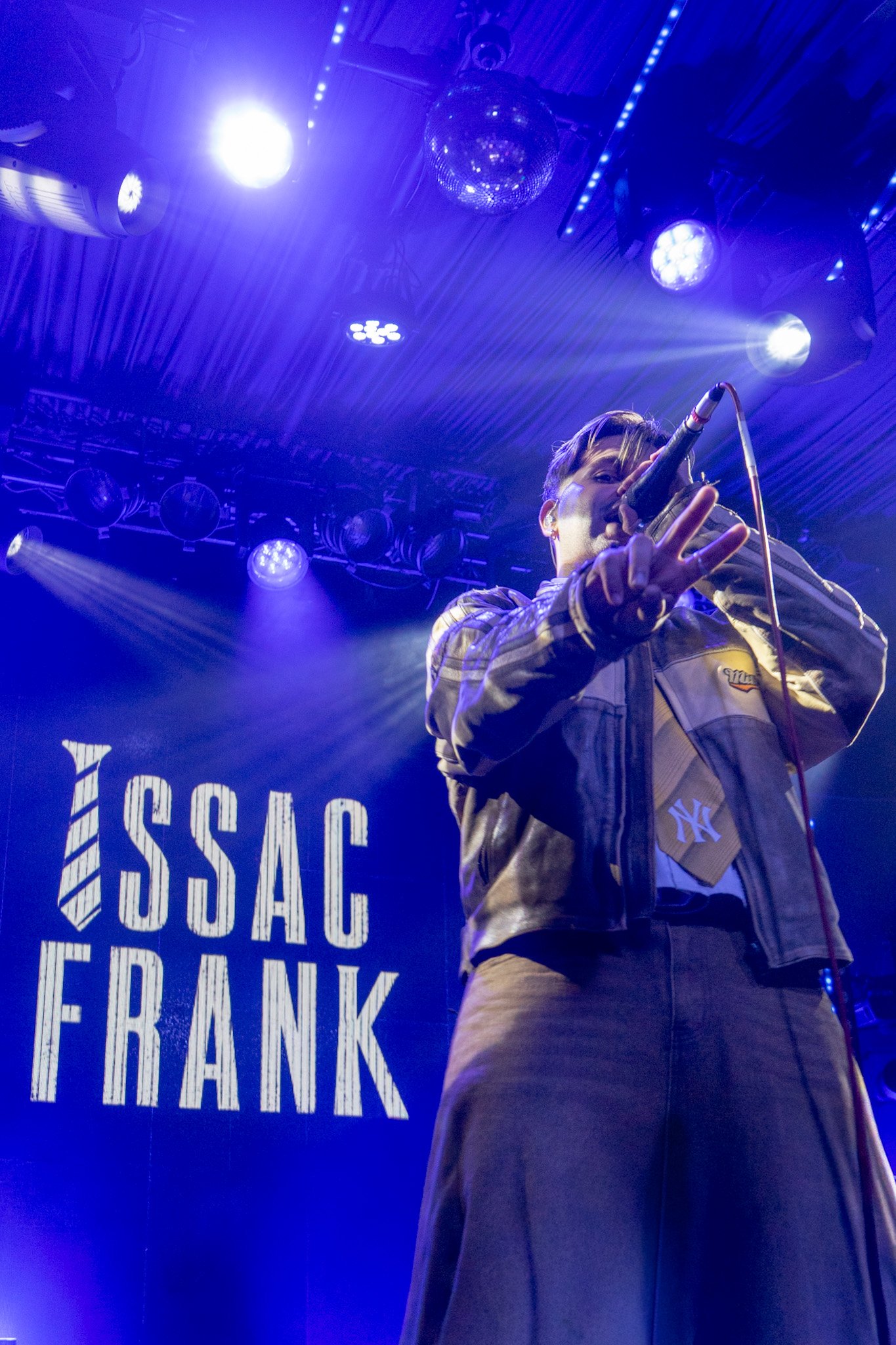Performing on stage with blue and purple lighting, a male singer with short hair wearing a beige jacket and brown pants.