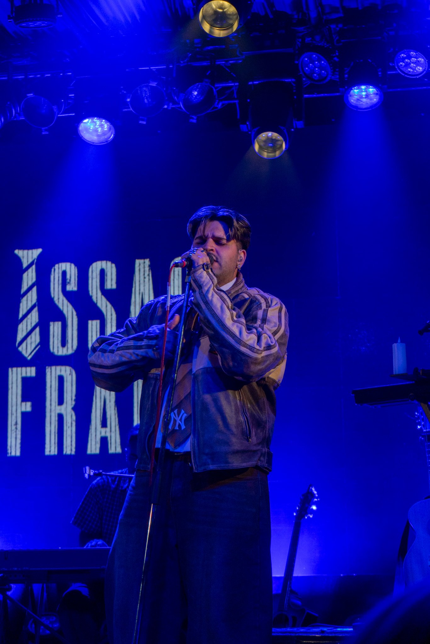 A young male singer with styled dark hair and earrings, wearing a plaid jacket and dark pants, performs on stage under blue lighting, holding a microphone close to his mouth.