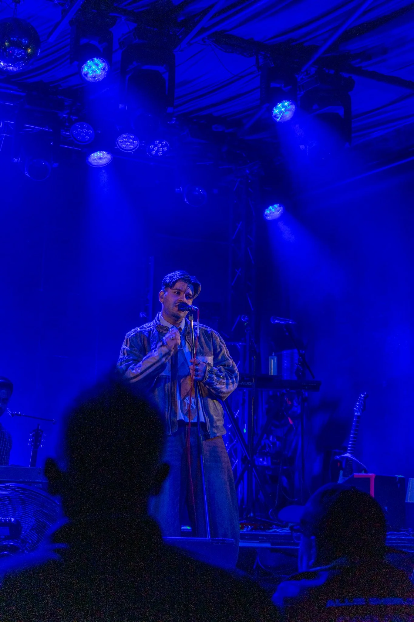 A male singer with a microphone on stage, illuminated by blue stage lights, with musical equipment and a partially visible audience in the background.