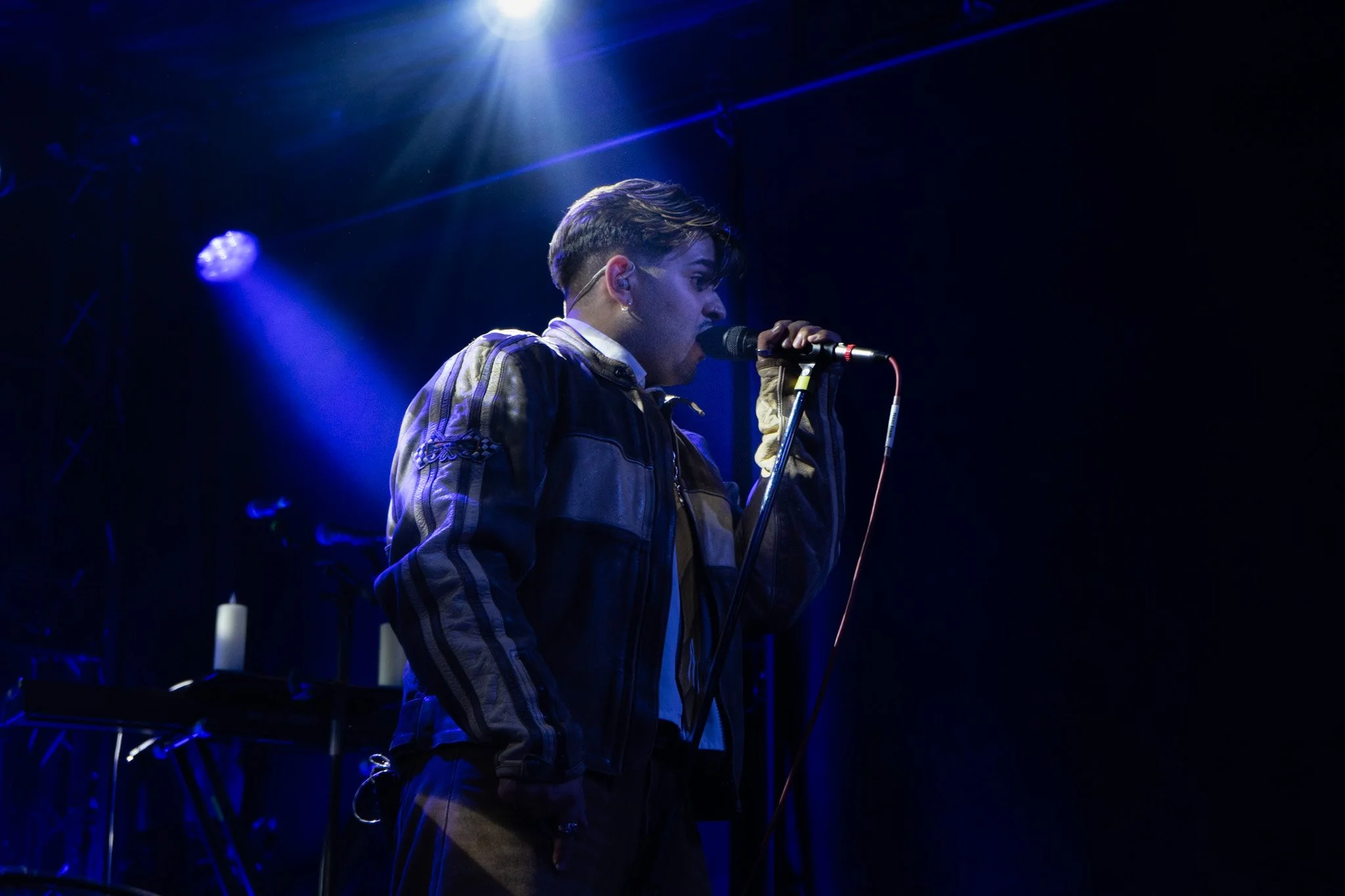 A young man singing into a microphone on stage, illuminated by a spotlight against a dark background.