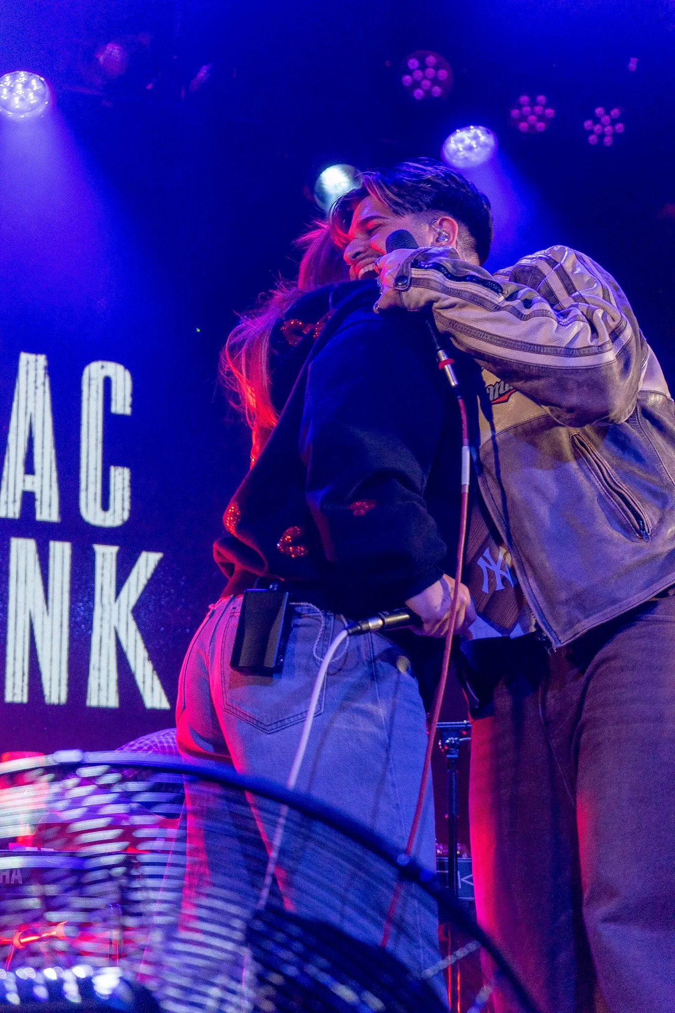 Two performers on stage sharing a hug, one holding a microphone. Stage lights and a large screen in the background.