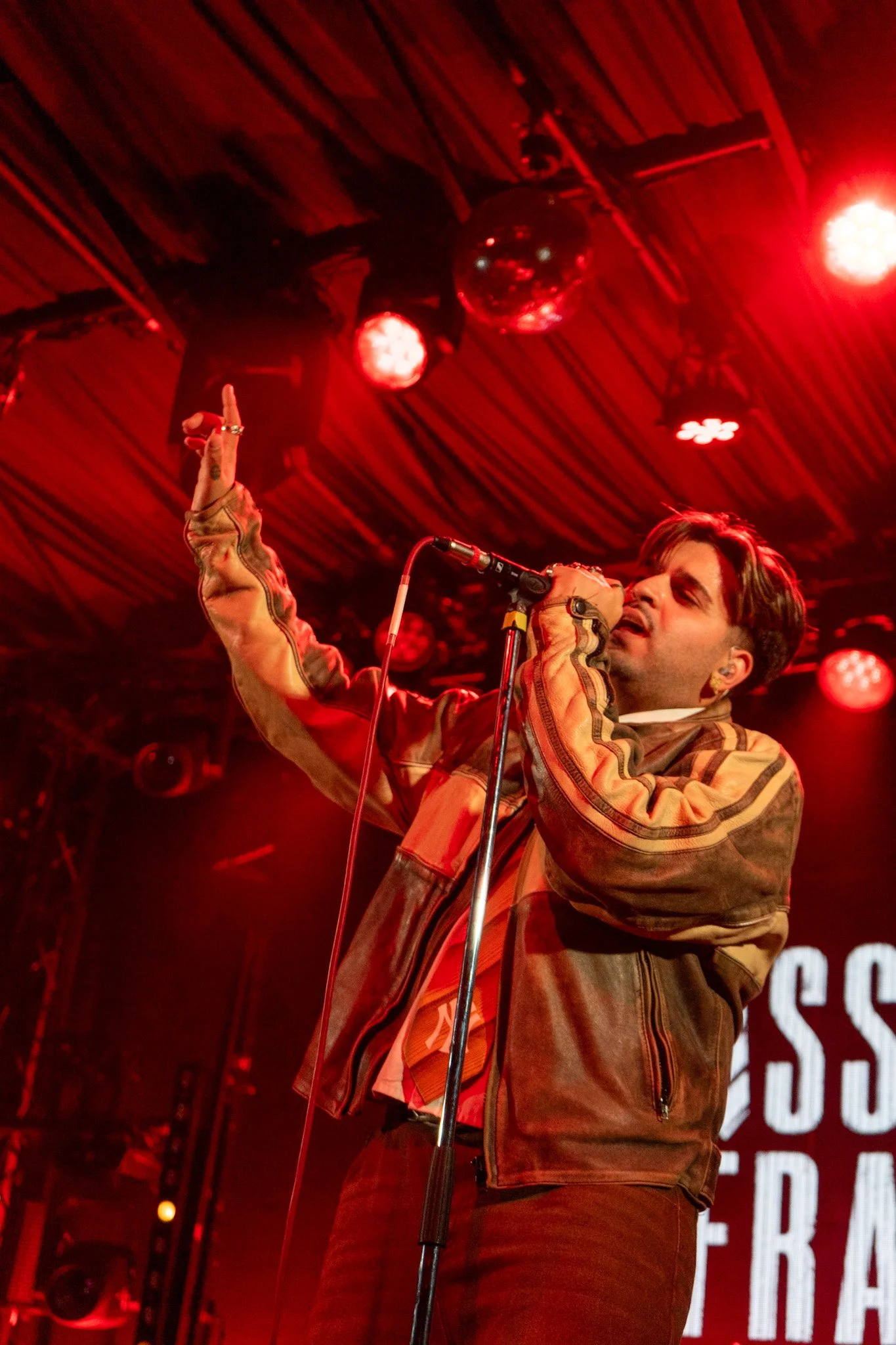 A male singer performing on stage in a brown leather jacket, holding a microphone, with red stage lights and a disco ball overhead.